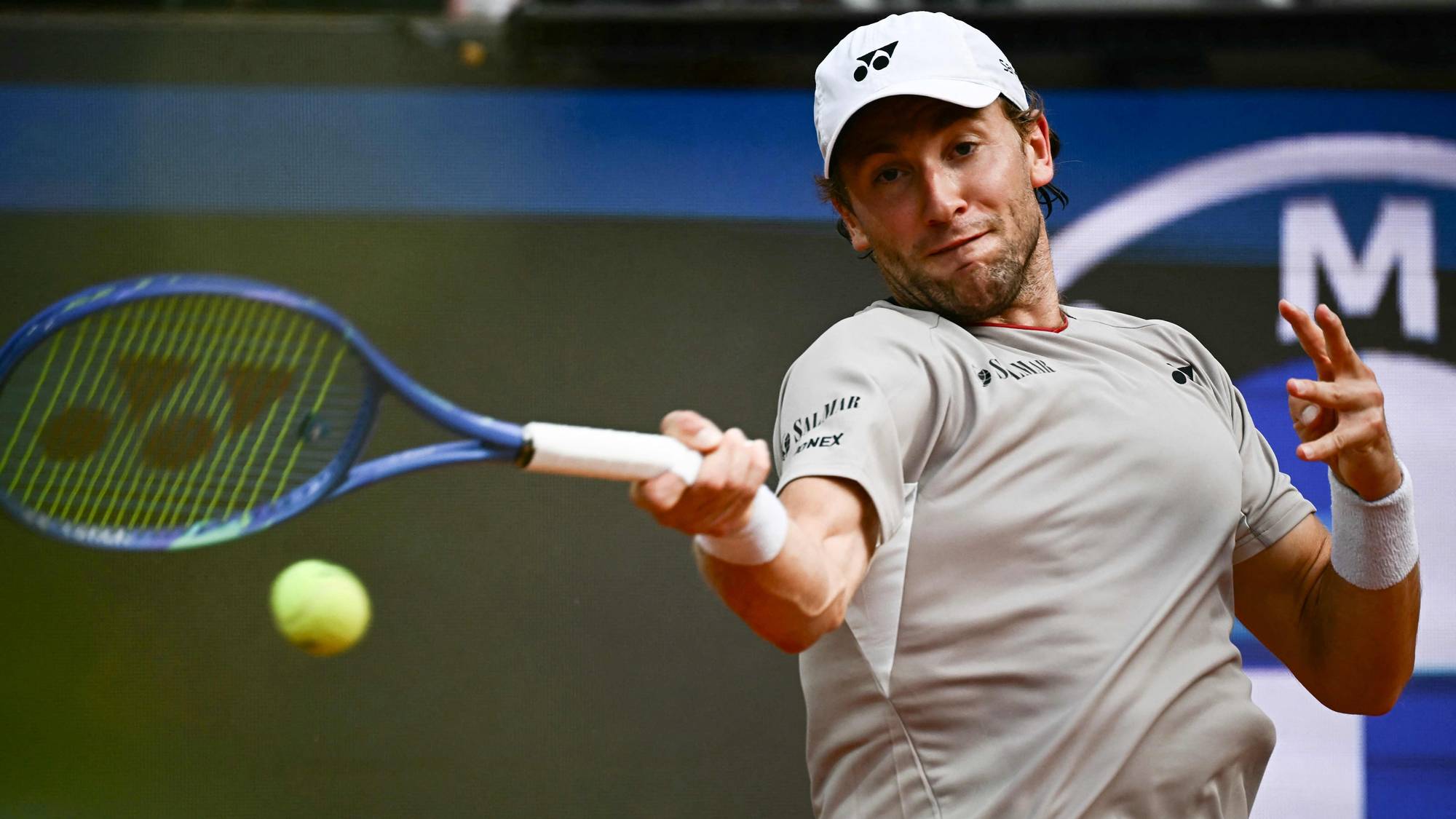 En mann i hvit caps og grå t-skjorte spiller tennis. Han holder en tennisracket i en slående bevegelse. Bak ham ser man et stort, uklart bilde. Det er en NTB-logo i midten av bildet. (Bildebeskrivelsen er laget av en KI-tjeneste)