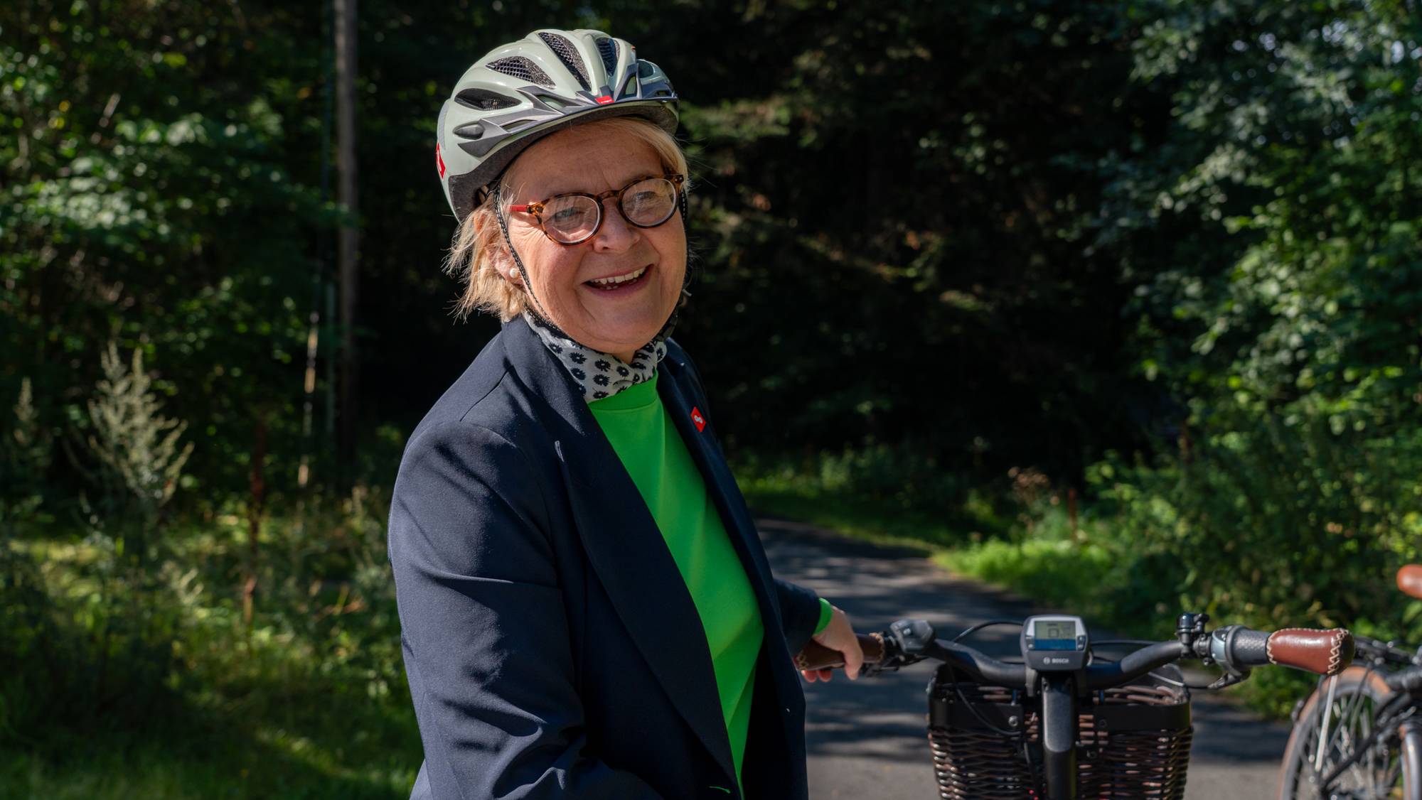 Kvinne med sykkelhjelm og briller smiler mens hun står ved en sykkel i skogsterreng.