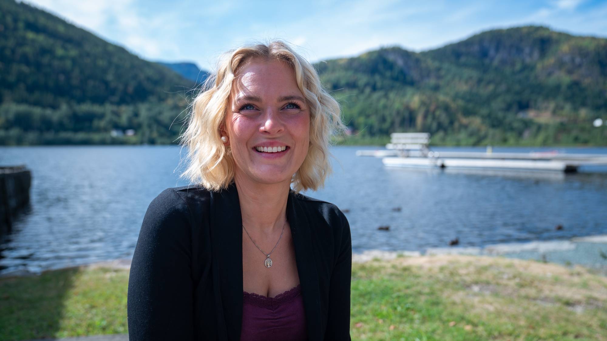 En kvinne sitter ved et vann og smiler.