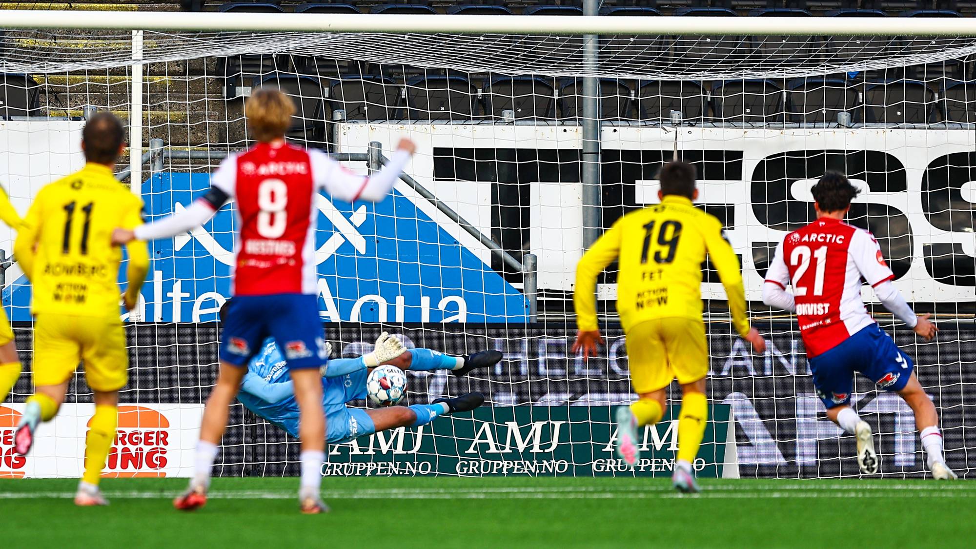 En keeper redder et skudd foran et mål. Fotballspillere i gule og røde drakter er på banen. Bak dem er tribuner og reklame. (Bildebeskrivelsen er laget av en KI-tjeneste)