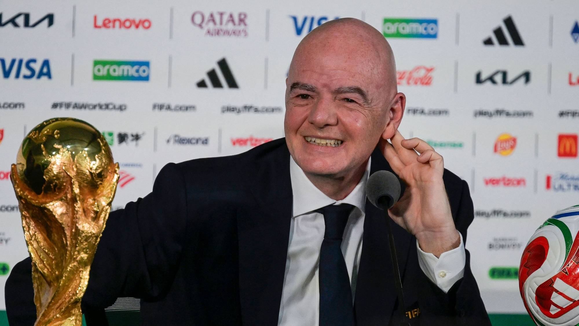 Gianni Infantino, president av FIFA, sitter bak et bord med fotball-VM-trofeet på sin venstre side. Infantino er iført en mørk dress og blått slips. En fotball ligger på bordet til høyre. Bak ham er en vegg med sponsorlogoer. (Bildebeskrivelsen er laget av en KI-tjeneste)