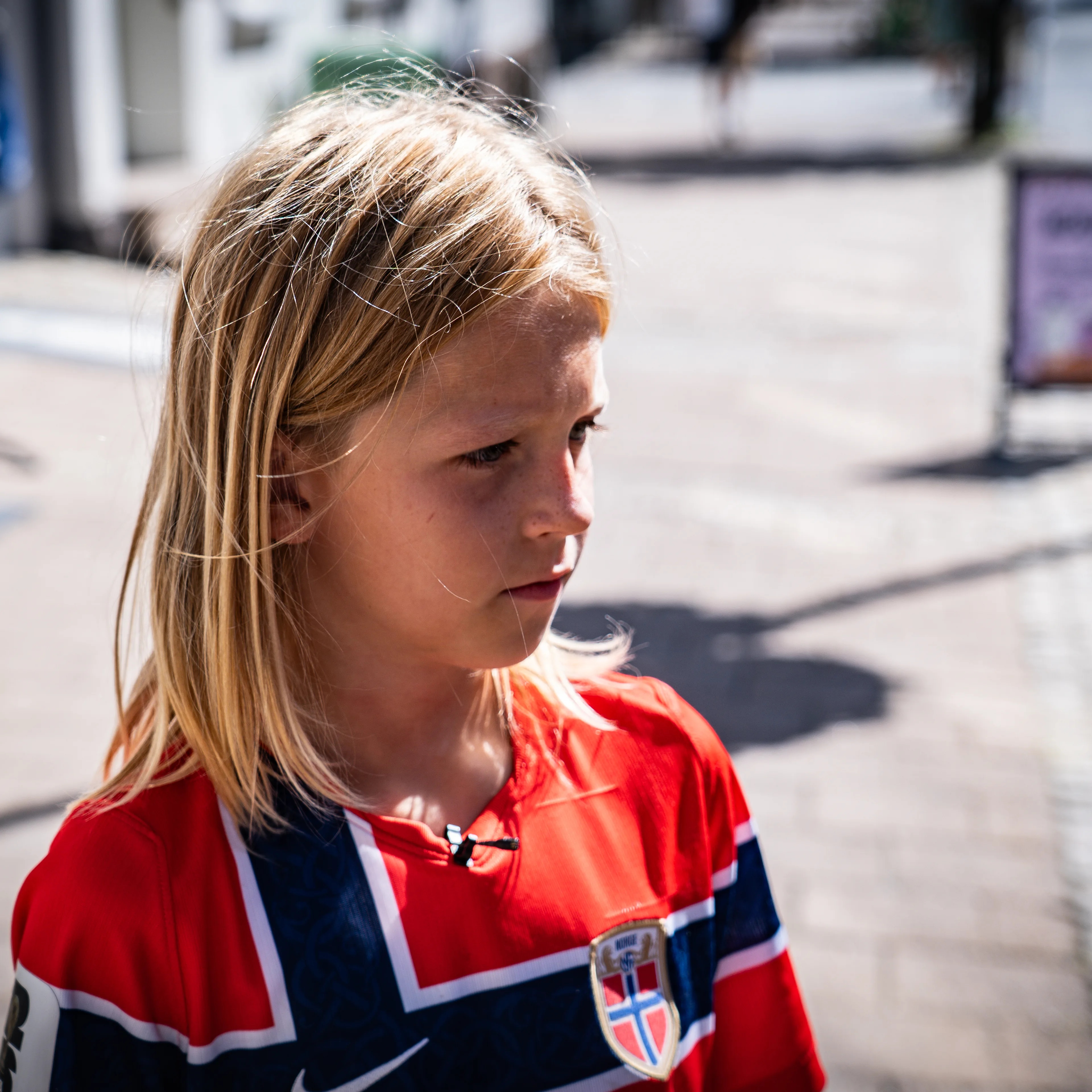 Barn med langt lyst hår i norsk landslagsdrakt står utendørs. (Bildebeskrivelsen er laget av en KI-tjeneste)