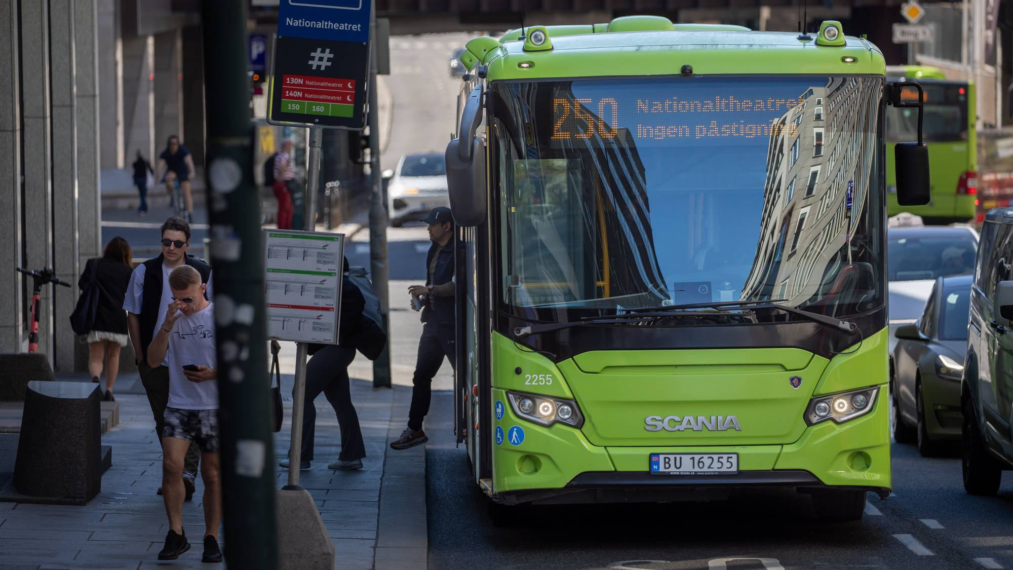 En grønn bybuss står ved en bussholdeplass. Flere personer venter og går på fortauet. Bussen har nummer 250 og skal til Nationaltheatret. Bakgrunnen viser bygninger og annen trafikk. (Bildebeskrivelsen er laget av en KI-tjeneste)