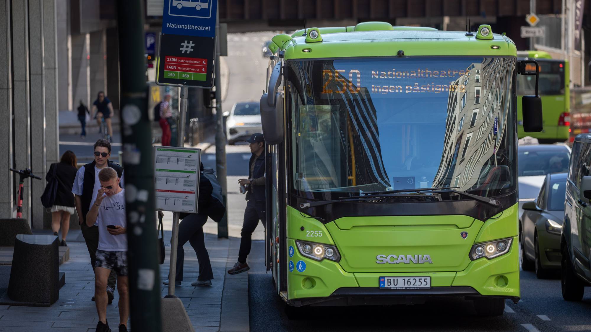 En grønn bybuss står ved en bussholdeplass. Flere personer venter og går på fortauet. Bussen har nummer 250 og skal til Nationaltheatret. Bakgrunnen viser bygninger og annen trafikk. (Bildebeskrivelsen er laget av en KI-tjeneste)