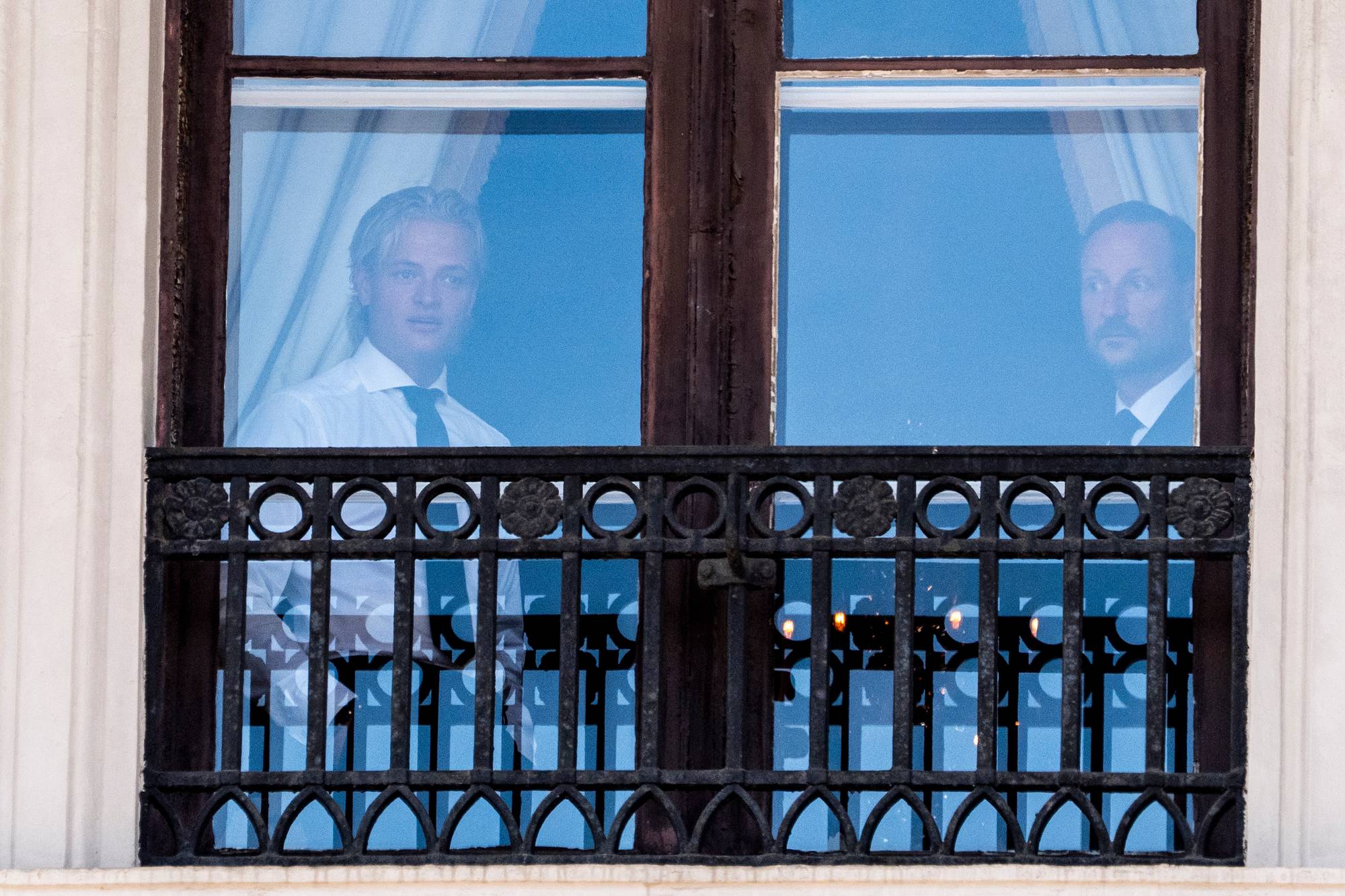 Kong Haakon og Marius Borg Høiby ser ut av et vindu med smijernsgelender. 