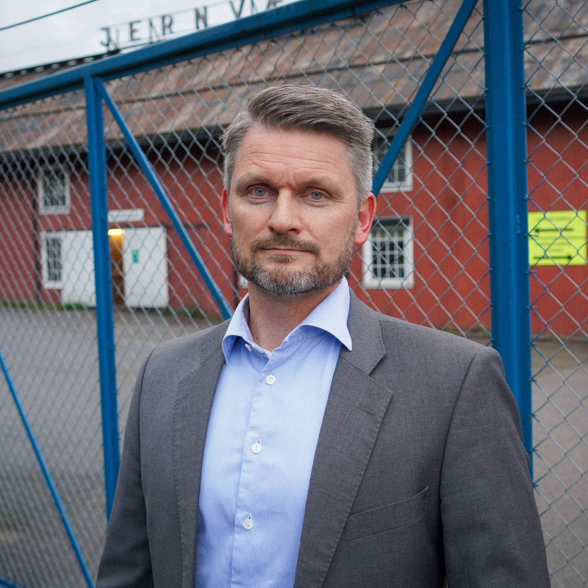 En mann i dress står foran et gjerde med en rød bygning i bakgrunnen. (Bildebeskrivelsen er laget av en KI-tjeneste)