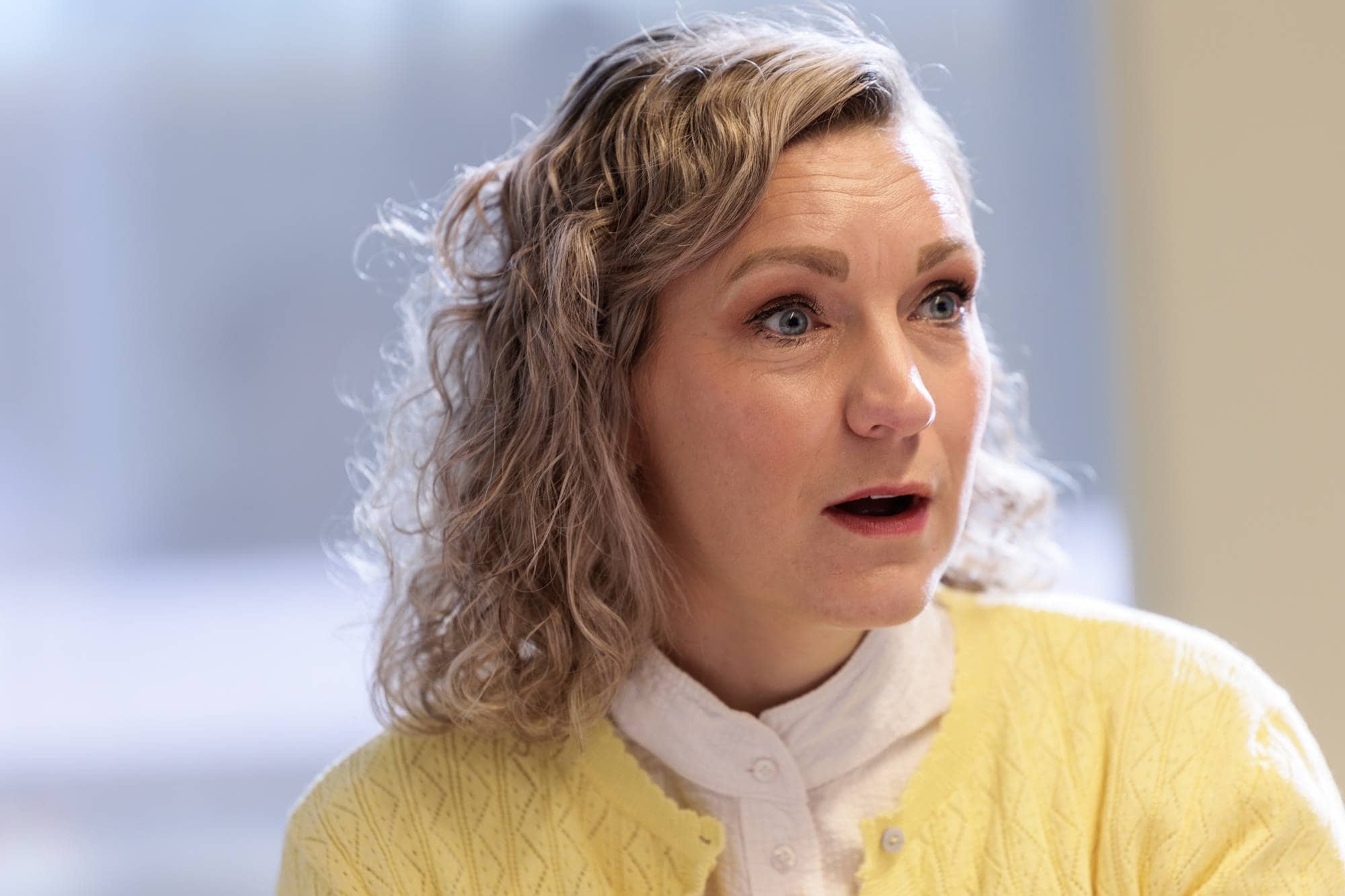 Kvinne med krøllete, blondt hår iført gul genser, sett i profil mens hun snakker. (Bildebeskrivelsen er laget av en KI-tjeneste)