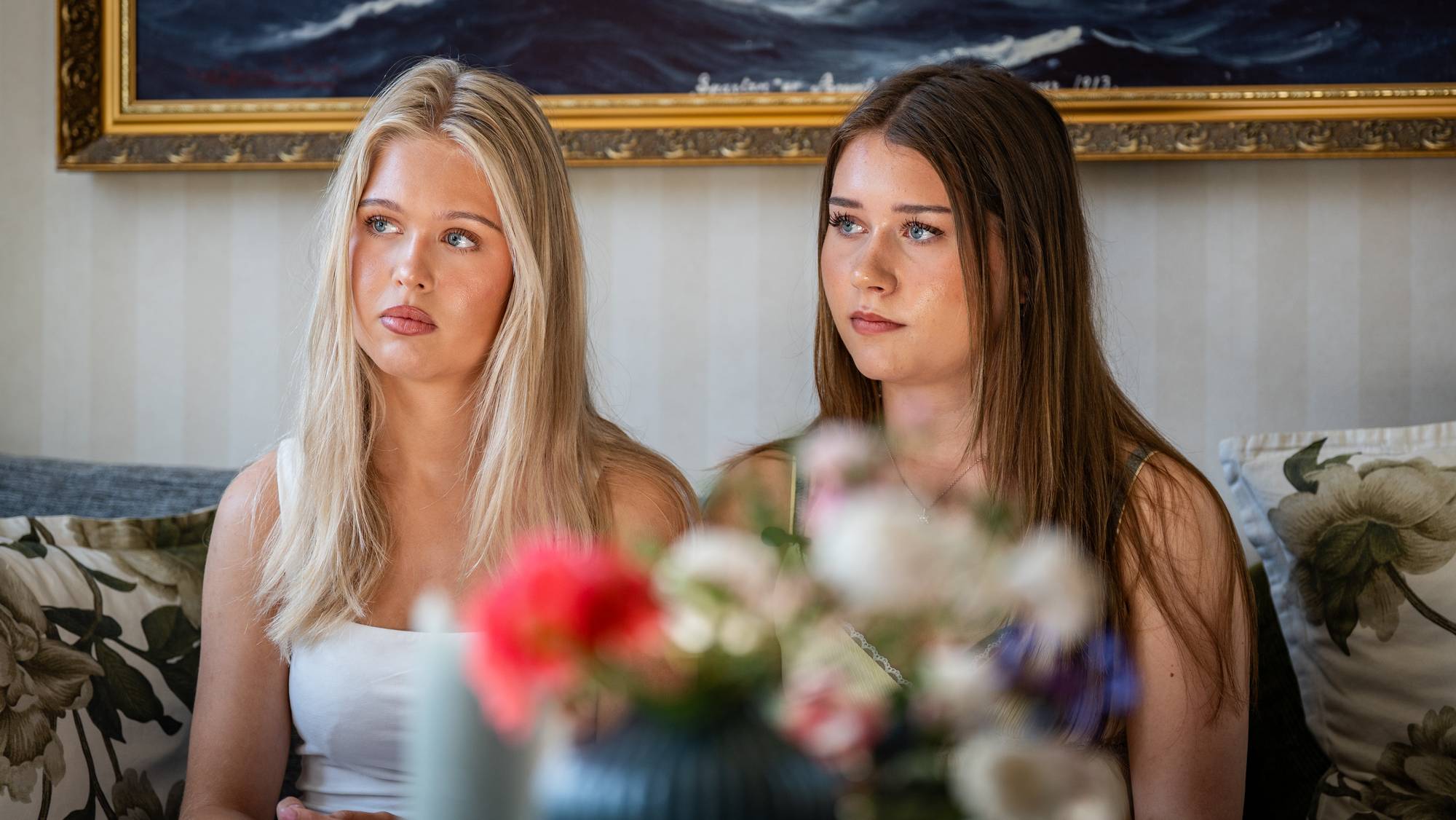 To unge kvinner sitter ved siden av hverandre i en sofa med maleri i bakgrunnen. (Bildebeskrivelsen er laget av en KI-tjeneste)
