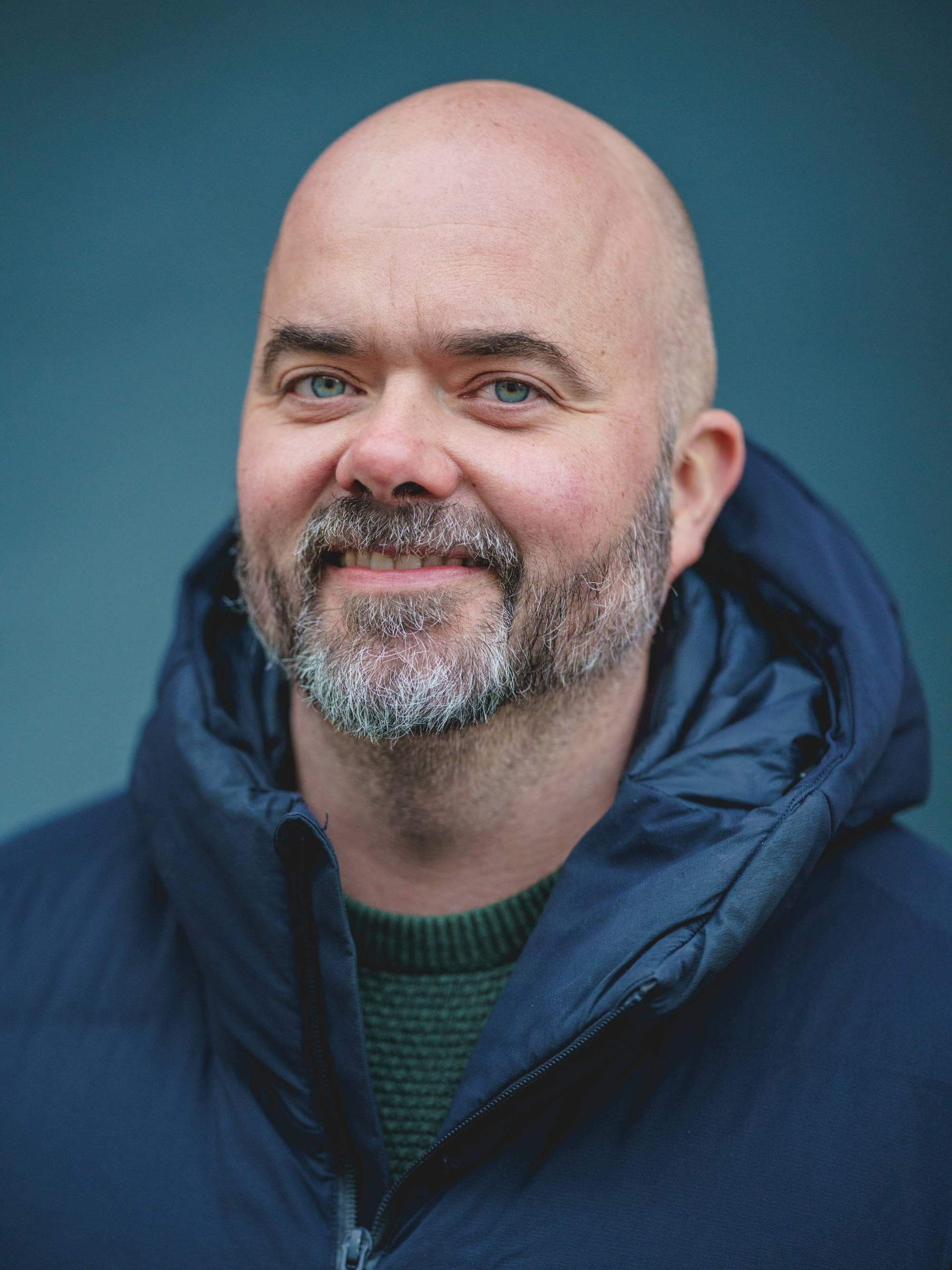 En mann med skallet hode og skjegg smiler mot kameraet. Han er iført en mørkeblå dunjakke over en grønn genser. Mannen er fotografert foran en blå bakgrunn. NTB-logoen er synlig over mannens bryst. (Bildebeskrivelsen er laget av en KI-tjeneste)