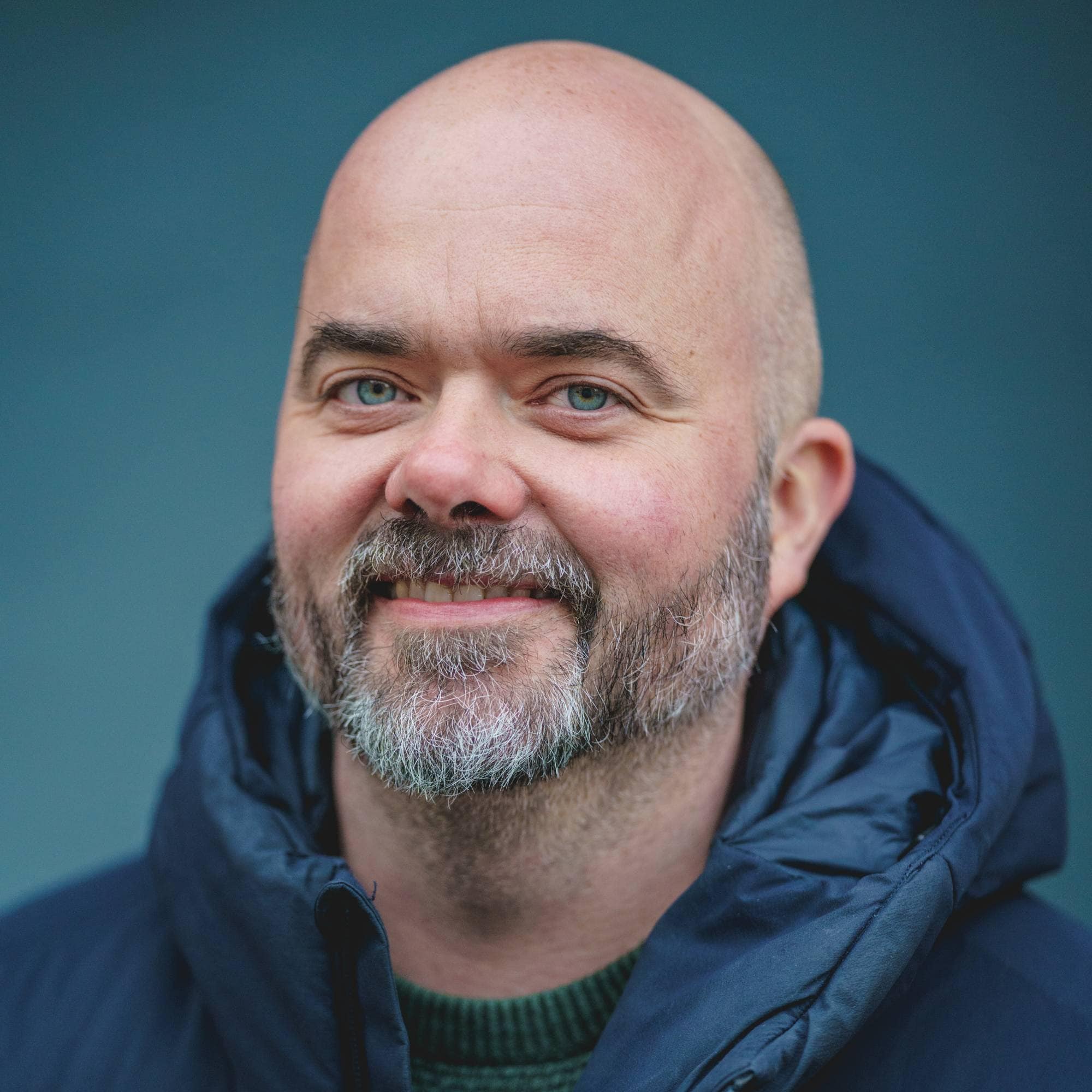 En mann med skallet hode og skjegg smiler mot kameraet. Han er iført en mørkeblå dunjakke over en grønn genser. Mannen er fotografert foran en blå bakgrunn. NTB-logoen er synlig over mannens bryst. (Bildebeskrivelsen er laget av en KI-tjeneste)