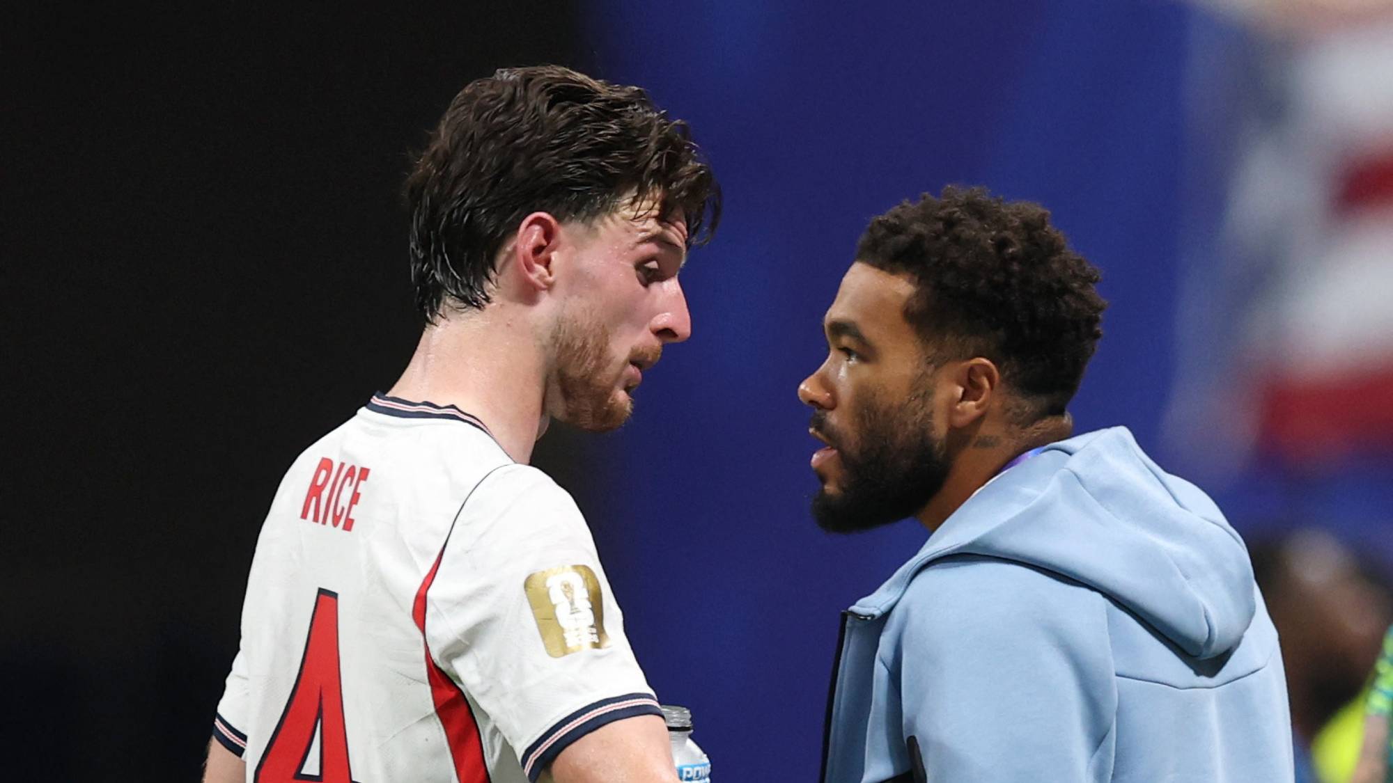Declan Rice i England-drakt snakker med en mann i lyseblå treningsdress på en fotballbane. (Bildebeskrivelsen er laget av en KI-tjeneste)