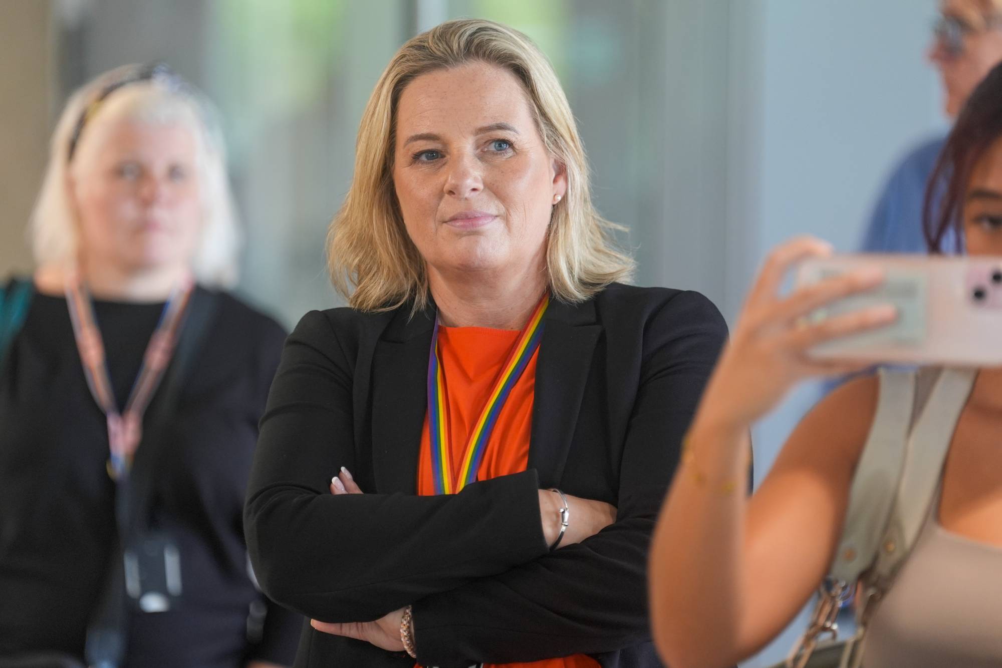 Anita Leirvik North med et regnbuefarget bånd rundt halsen. Andre personer er delvis synlige i bakgrunnen. (Bildebeskrivelsen er laget av en KI-tjeneste)