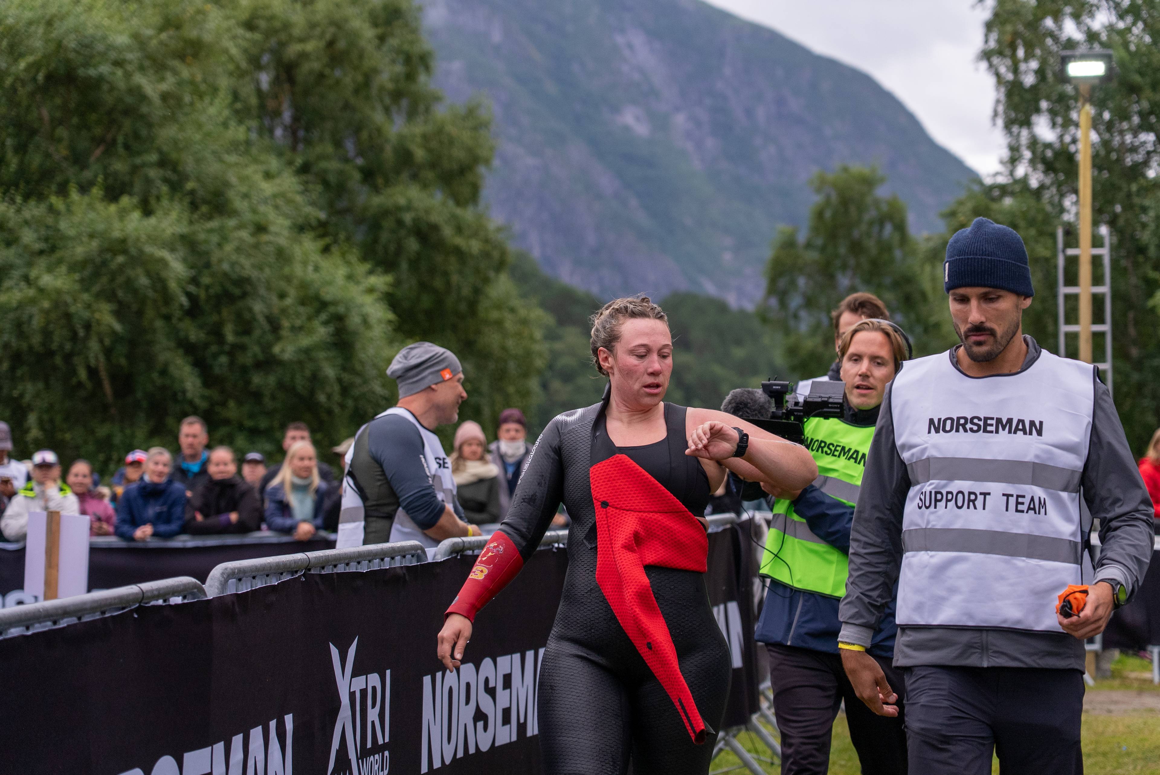 Triatlet i våtdrakt sjekker klokken ved målområdet til Norseman med støtteteam i bakgrunnen. (Bildebeskrivelsen er laget av en KI-tjeneste)