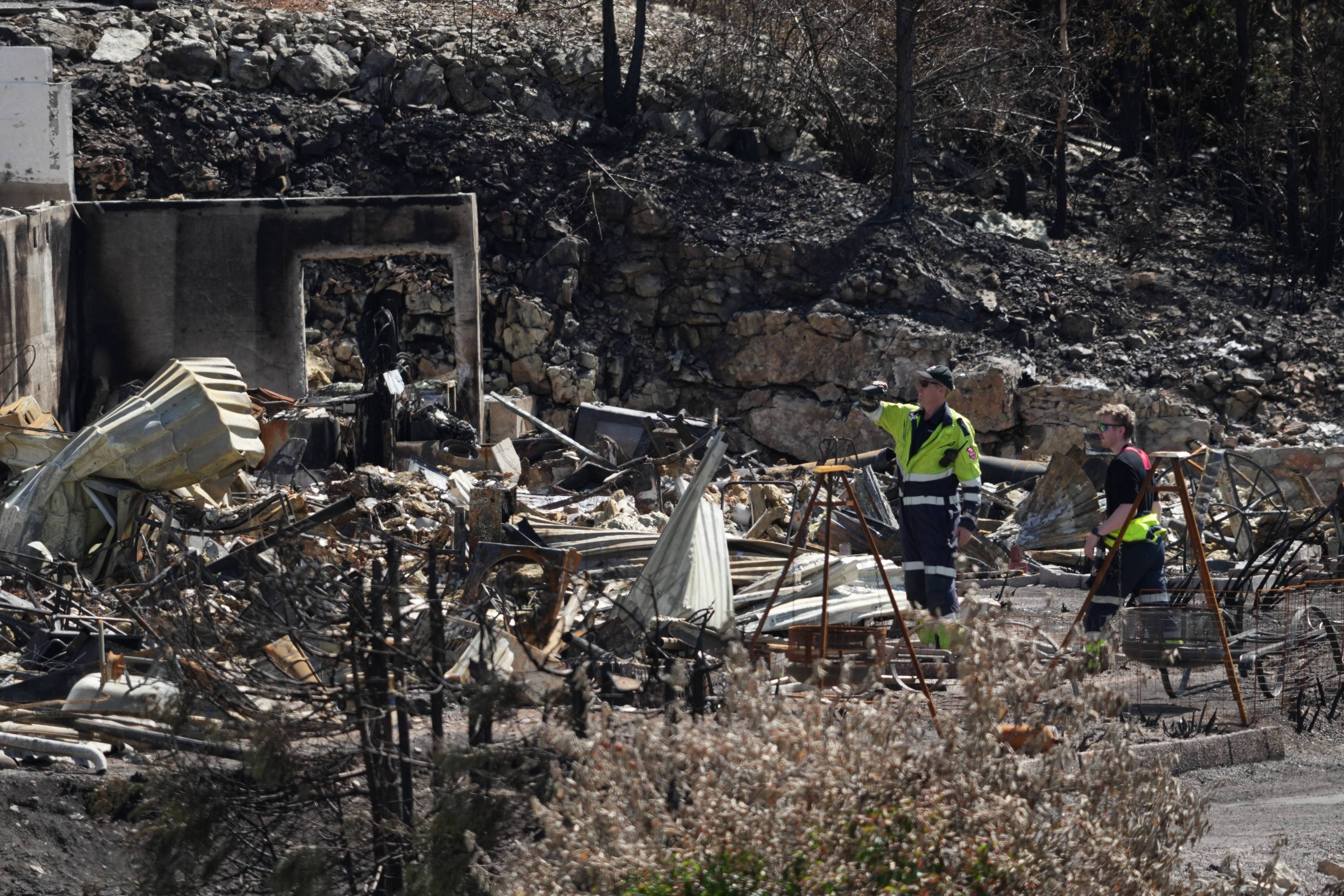 To personer undersøker restene av et nedbrent hus i et område preget av skogbrann. (Bildebeskrivelsen er laget av en KI-tjeneste)