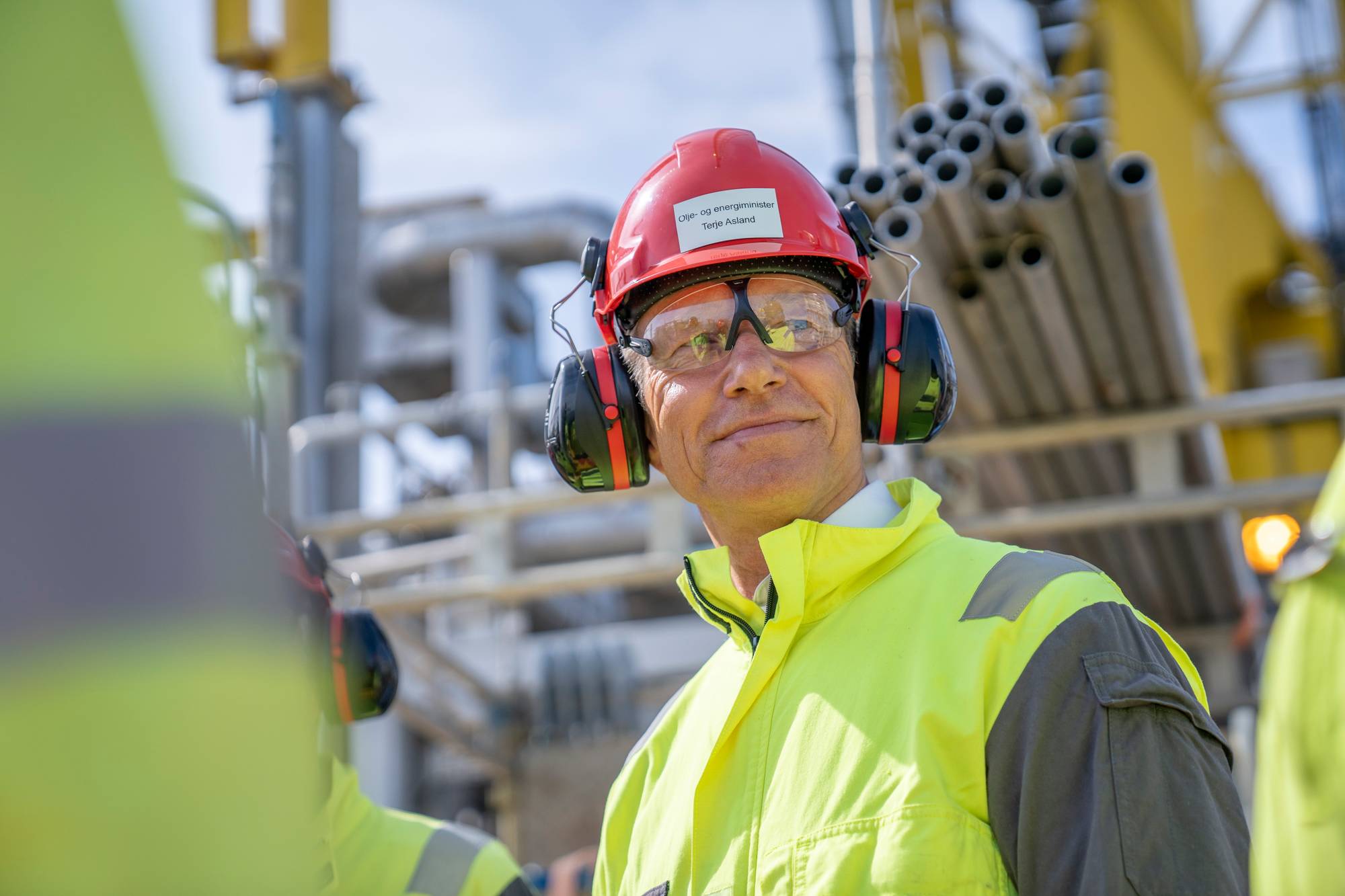 Olje- og energiminister Terje Aasland på plattforma Njord i Norskehavet. Han har på seg ein refleksfarga kjeledress og har ein raud hjelm med øyreklokker på seg. Bak han ser vi boreriggen.