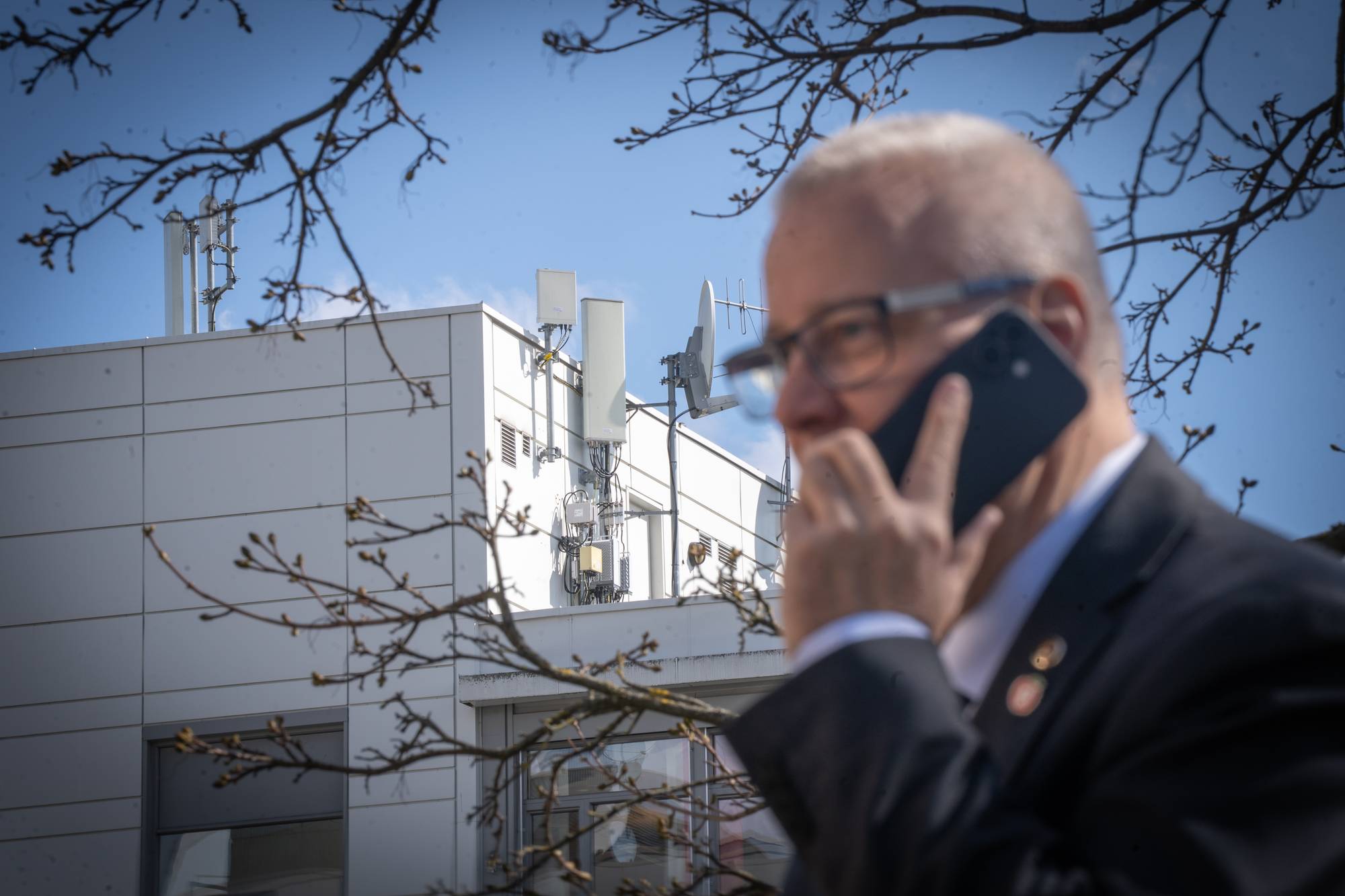 En mann i dress snakker i en mobiltelefon. I bakgrunnen er en bygning med antenner på taket. En gren med knopper henger foran. (Bildebeskrivelsen er laget av en KI-tjeneste)