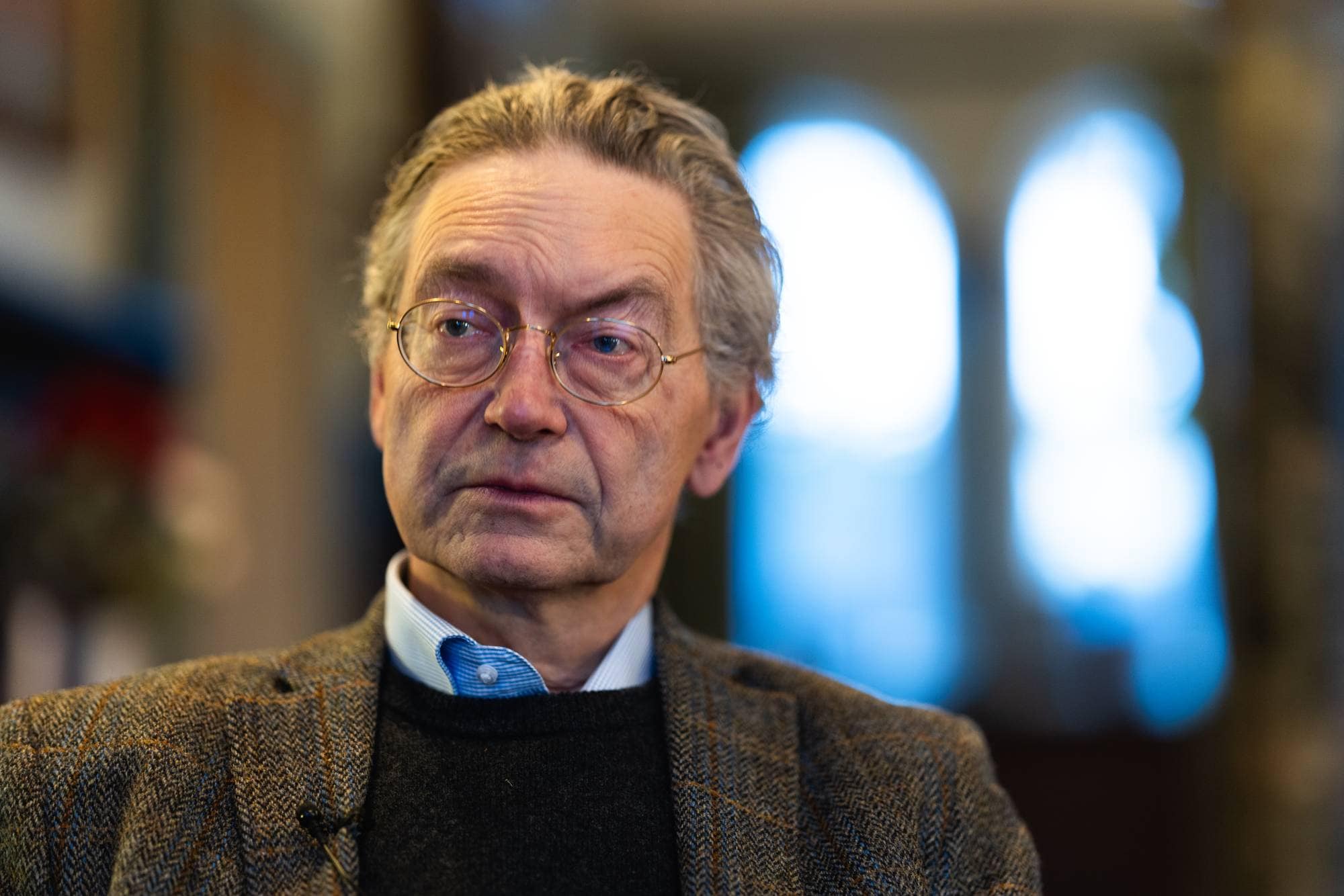 En mann sitter i en stol, iført en jakke med skråstripet mønster. Han har på seg en svart genser og en blå skjorte. Bakgrunnen viser svake konturer av innendørs interiør med uklare lys. Belysningen gir en dypere atmosfære til bildet. 