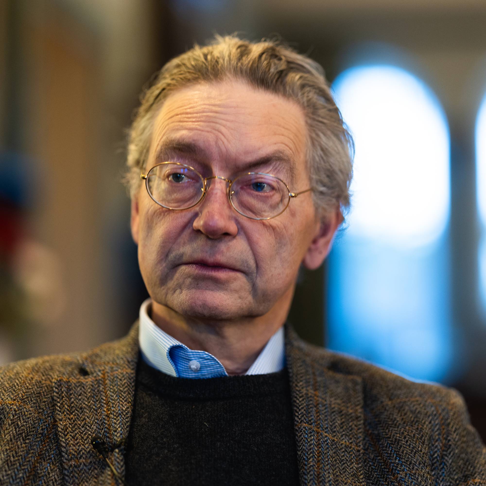 En mann sitter i en stol, iført en jakke med skråstripet mønster. Han har på seg en svart genser og en blå skjorte. Bakgrunnen viser svake konturer av innendørs interiør med uklare lys. Belysningen gir en dypere atmosfære til bildet. 