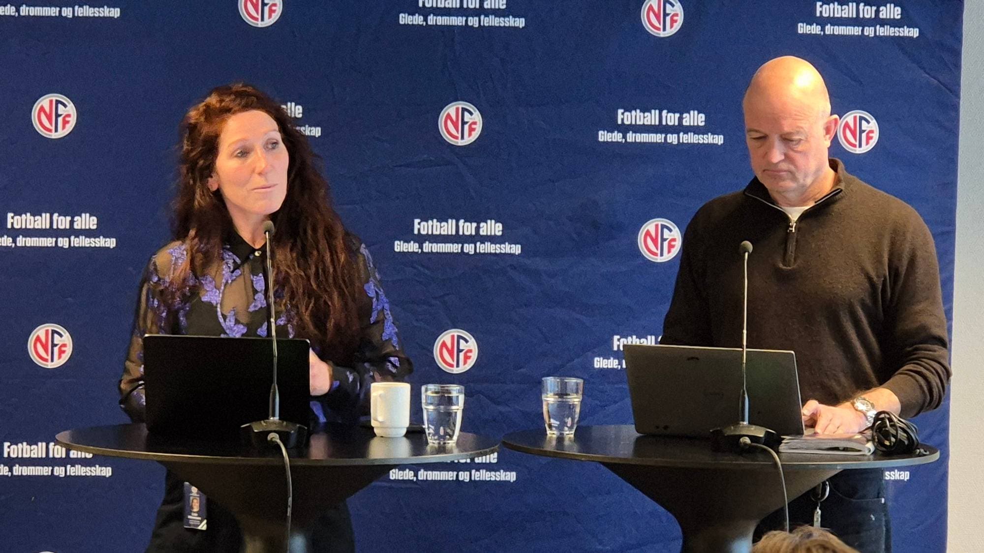 En kvinne og en mann står bak et podium foran en blå bakgrunn med Norges Fotballforbunds logo. Kvinnen har en bærbar datamaskin foran seg på podiumet. Mannen står ved siden av et podium med en bærbar datamaskin foran seg. Flere personer sitter på stoler i forgrunnen, vendt bort fra kamera. (Bildebeskrivelsen er laget av en KI-tjeneste)