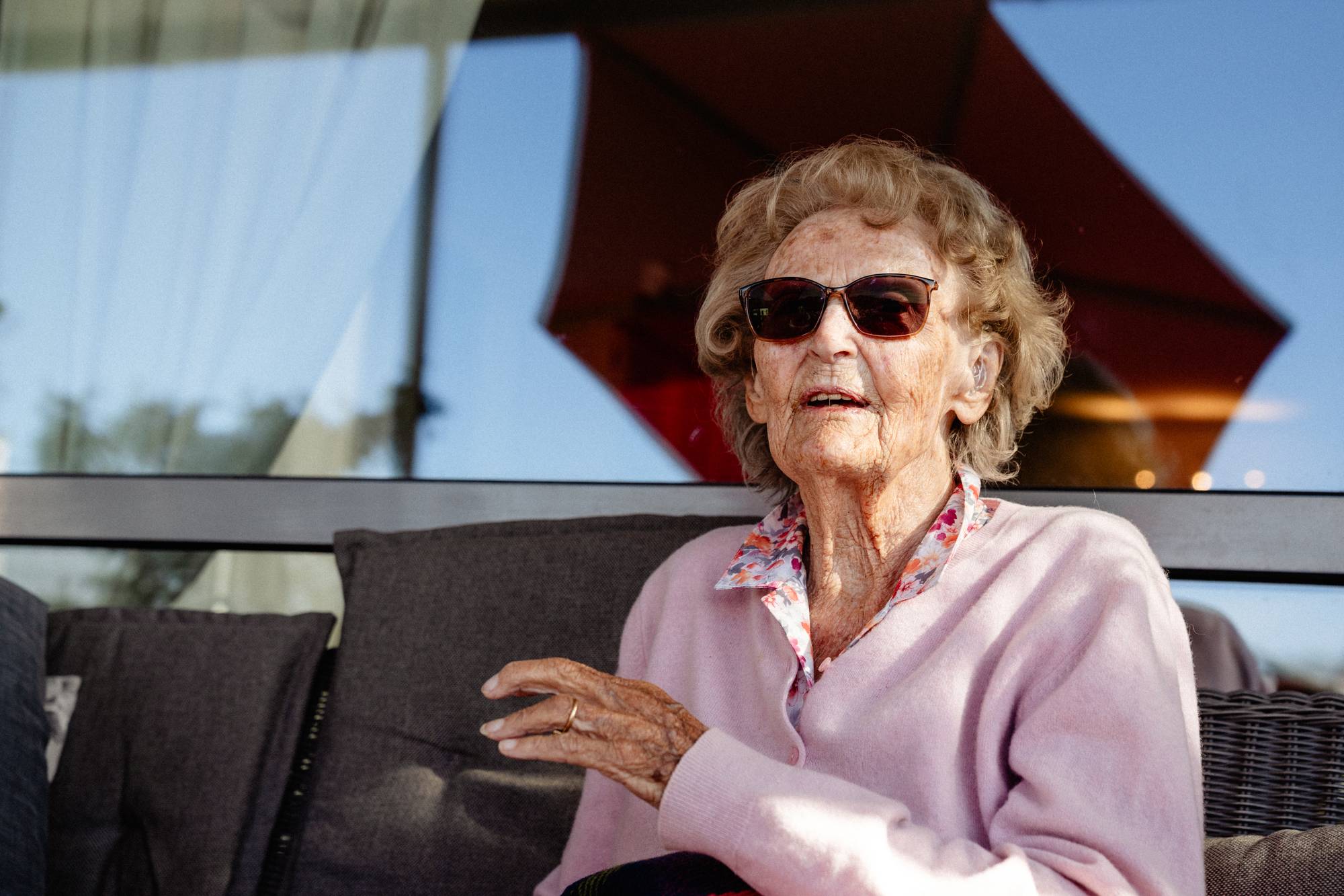 Eldre kvinne med solbriller sitter ute og snakker. Hun har på seg en lyserosa cardigan. (Bildebeskrivelsen er laget av en KI-tjeneste)