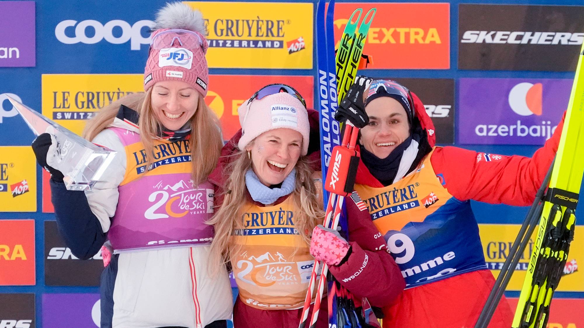 Tre glade kvinnelige langrennsløpere står på pallen foran en reklamevegg etter Tour de Ski. (Bildebeskrivelsen er laget av en KI-tjeneste)