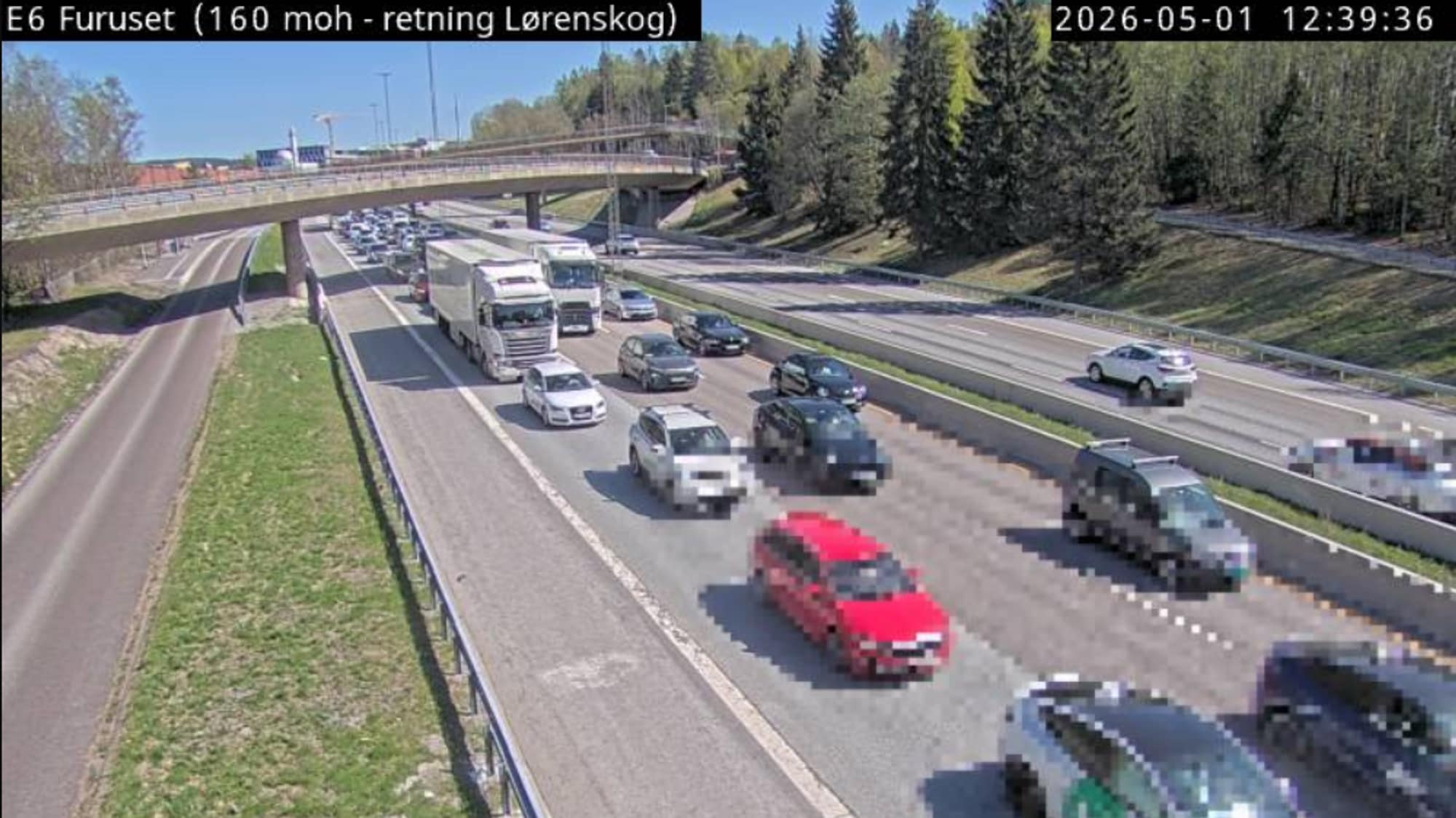 Trafikk på E6 ved Furuset, i retning Lørenskog. Mange biler og noen lastebiler står i kø på motorveien. Overkjøringsbro og trær langs veien. Trafikken ser ut til å være tett på ettermiddagen. (Bildebeskrivelsen er laget av en KI-tjeneste)