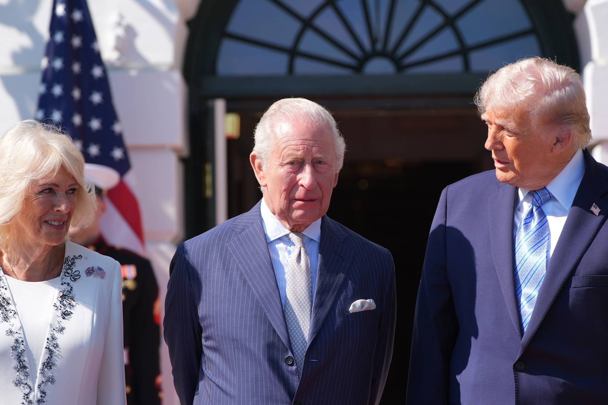 Prins Charles og Donald Trump står sammen foran en bygning. Camilla Parker Bowles står til venstre for dem. En amerikansk flagg er synlig i bakgrunnen. (Bildebeskrivelsen er laget av en KI-tjeneste)