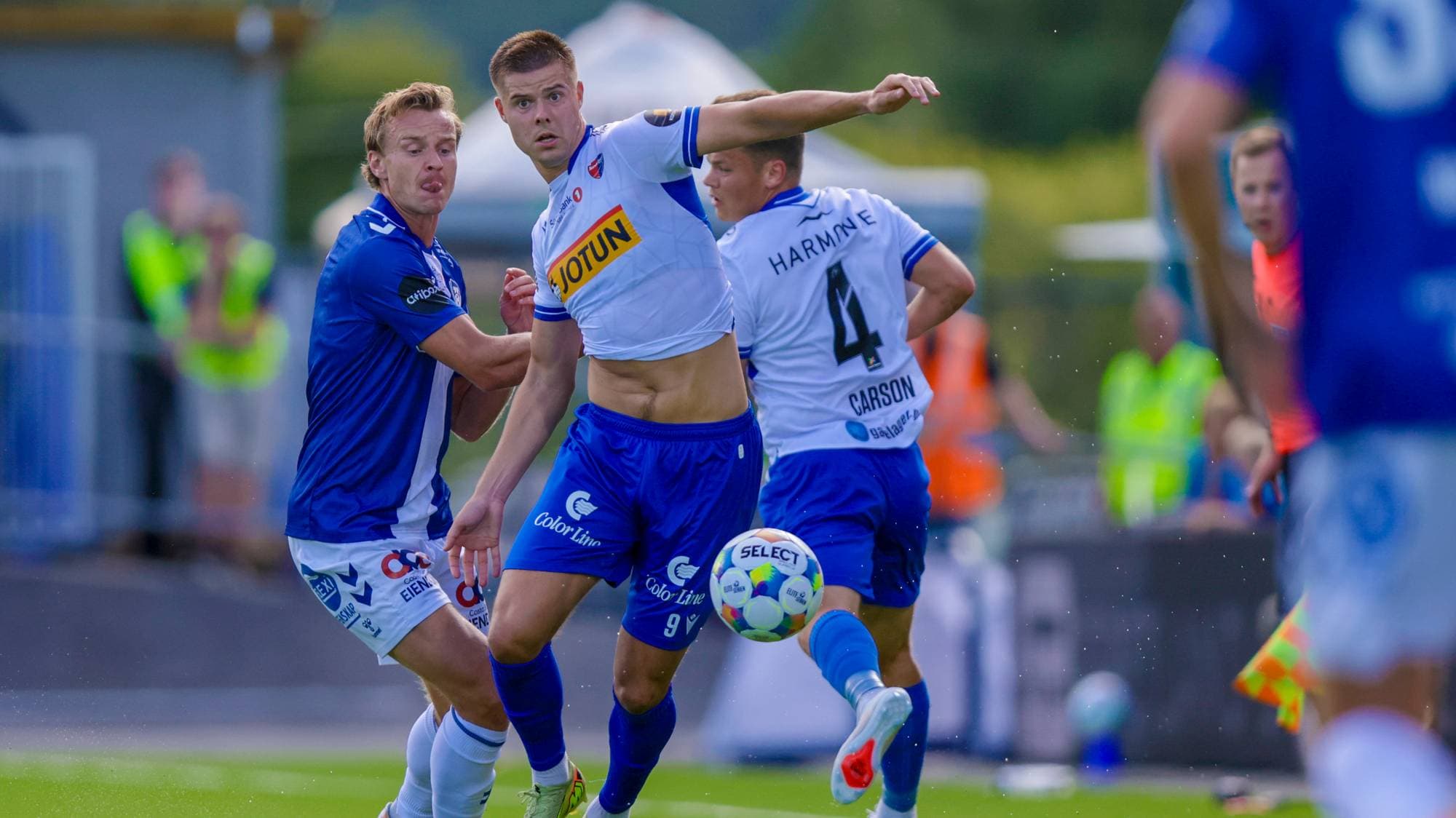 Fotballspillere i duell om ballen under en kamp på en gressbane. (Bildebeskrivelsen er laget av en KI-tjeneste)