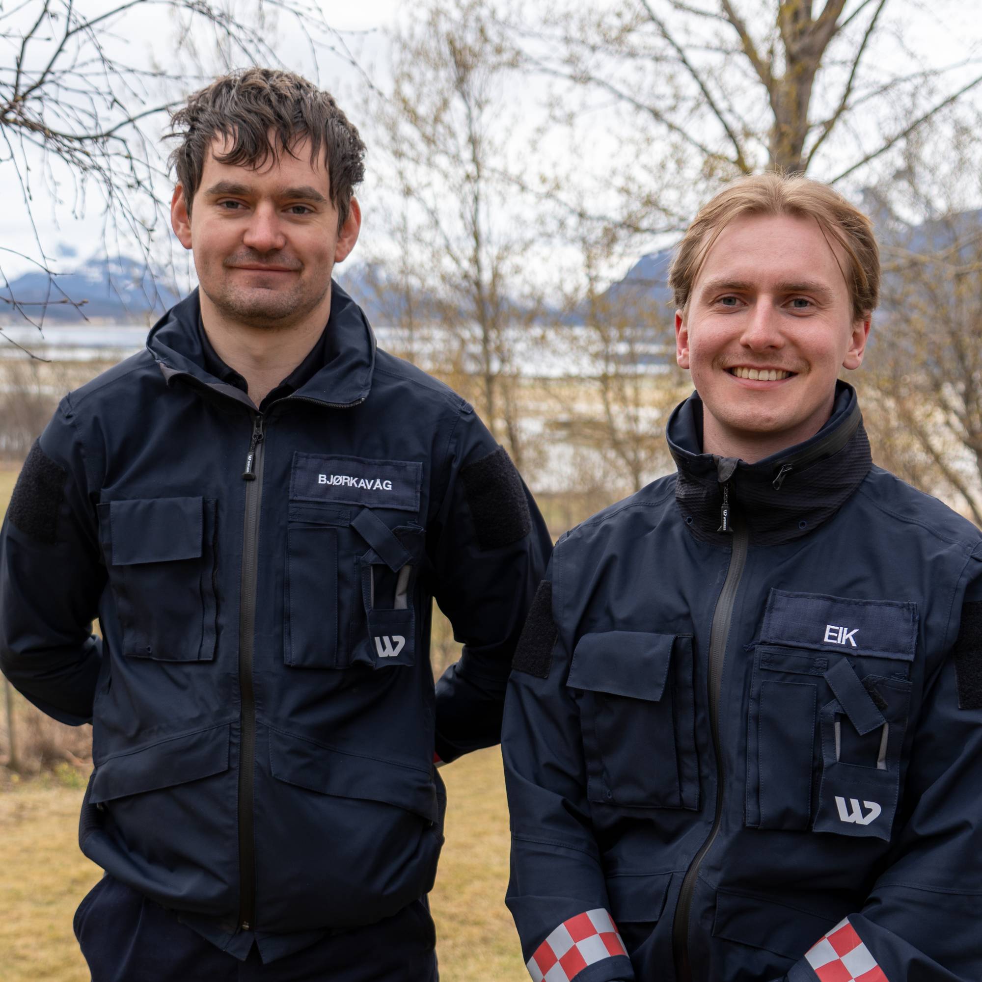 To menn i mørkeblå uniform står utendørs med skog og fjell i bakgrunnen. Den ene mannen har et navneskilt med "BJØRKAVAG" på jakken sin. Den andre mannen har et navneskilt med "EIK" på jakken sin. Begge ser mot kamera og smiler. (Bildebeskrivelsen er laget av en KI-tjeneste)