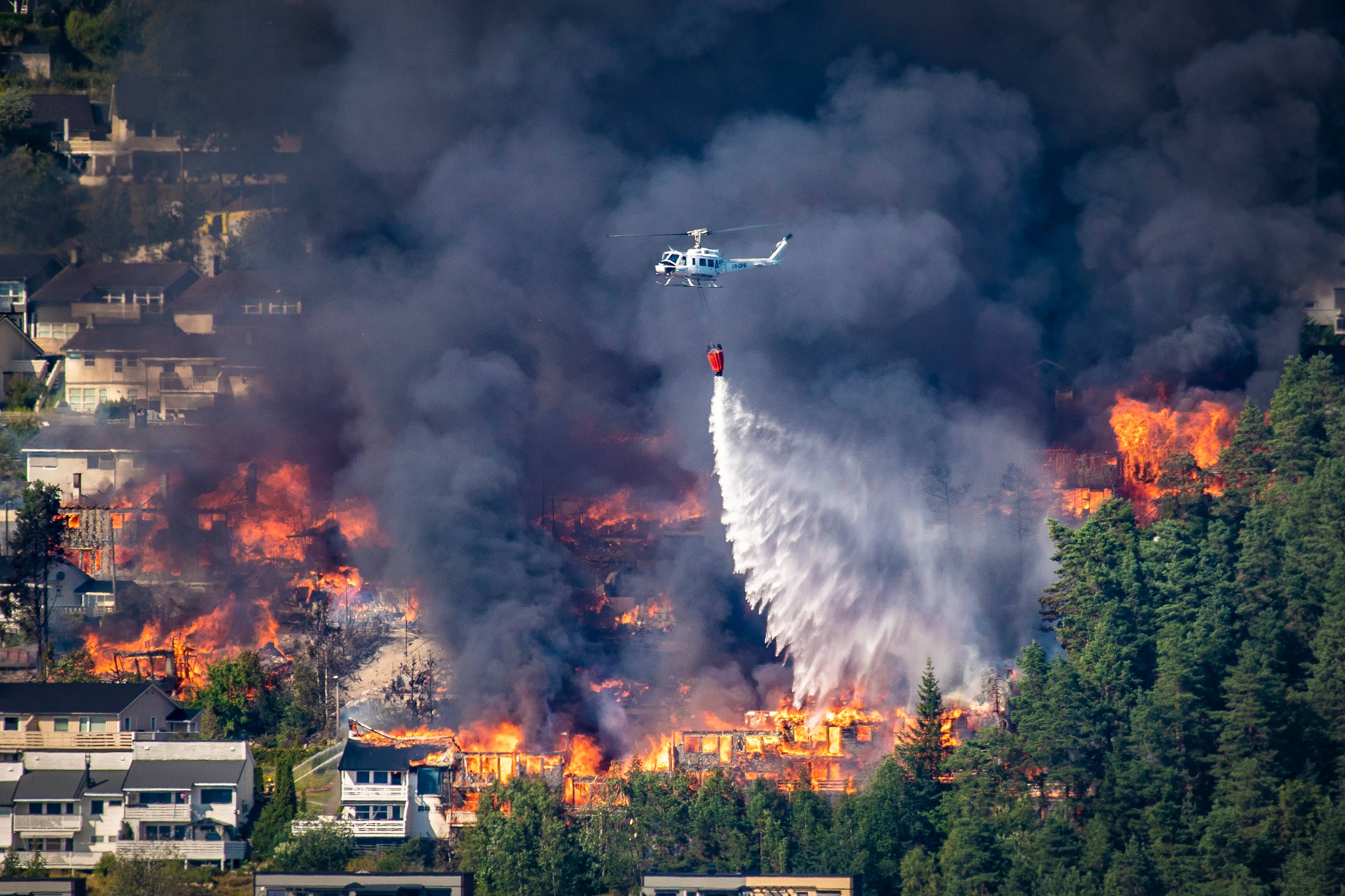 Et helikopter slipper vann over en boligbrann i et skogsområde omgitt av røyk. (Bildebeskrivelsen er laget av en KI-tjeneste)