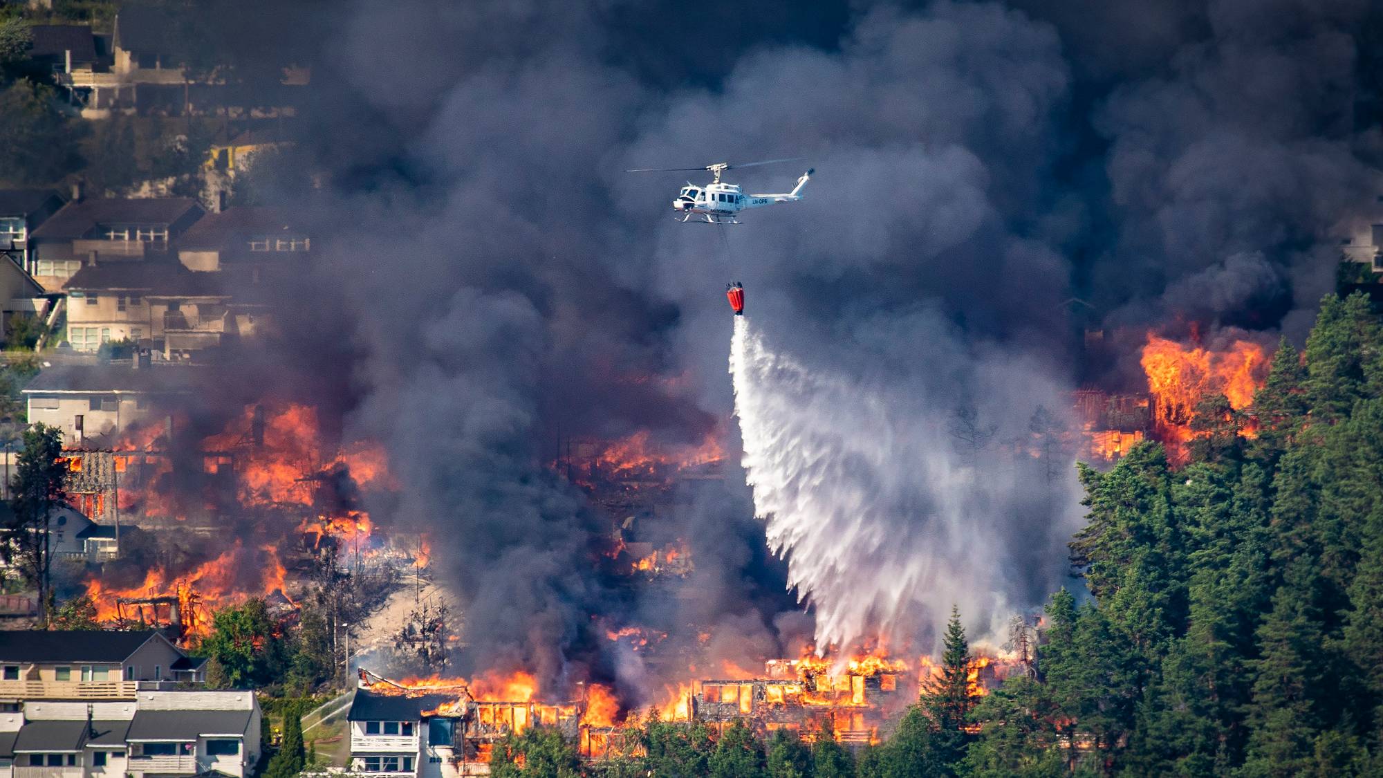 Et helikopter slipper vann over en boligbrann i et skogsområde omgitt av røyk. (Bildebeskrivelsen er laget av en KI-tjeneste)