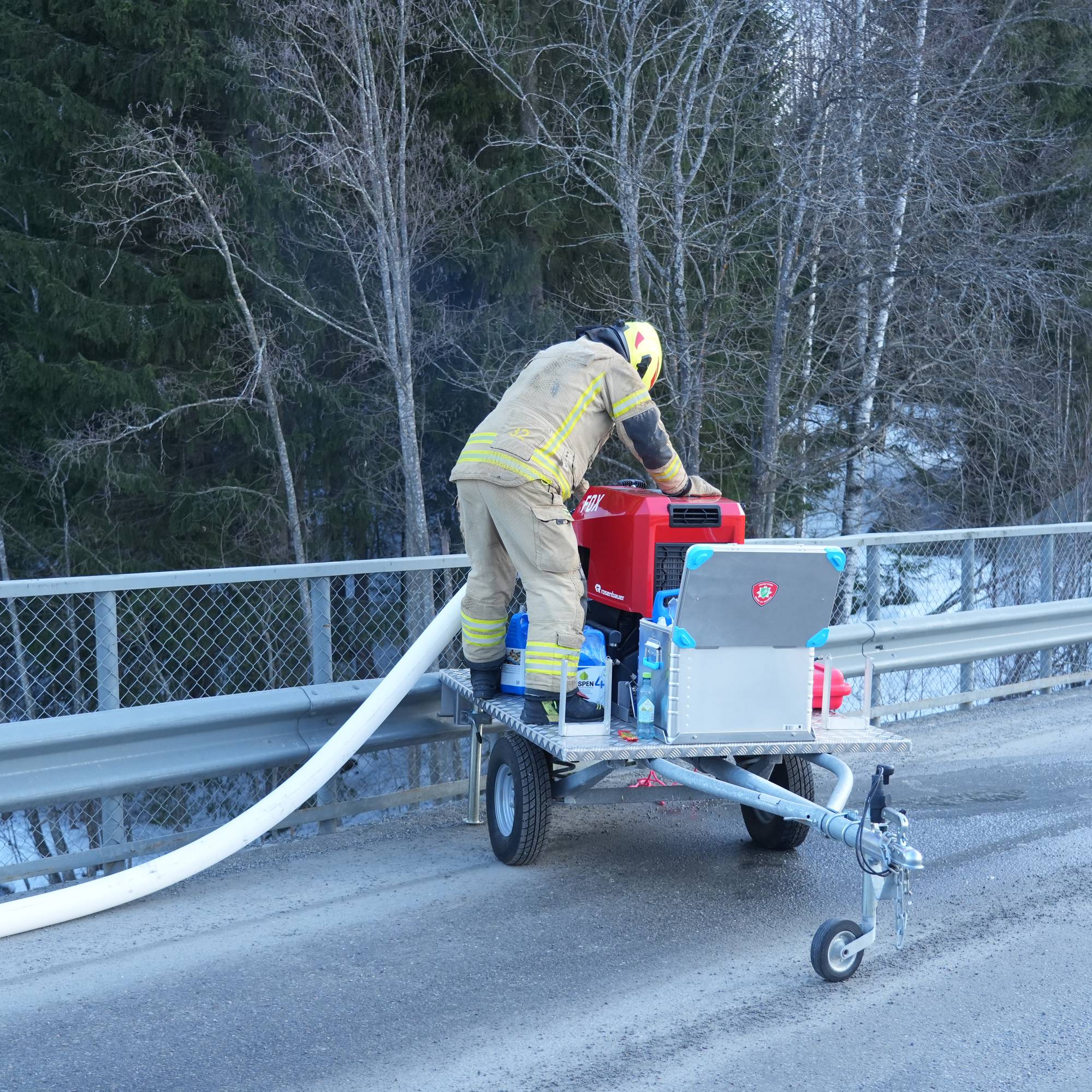 En brannmann i uniform arbeider med utstyr på en tilhenger på en bruflate. Han justerer en rød motor, mens en hvit slange strekker seg over kanten av brua. Omgivelsene består av trær og snø, og en del av veien er synlig. Bildet viser et situasjonsbilde fra en brannvernsoperasjon. (Bildebeskrivelsen er laget av en KI-tjeneste)