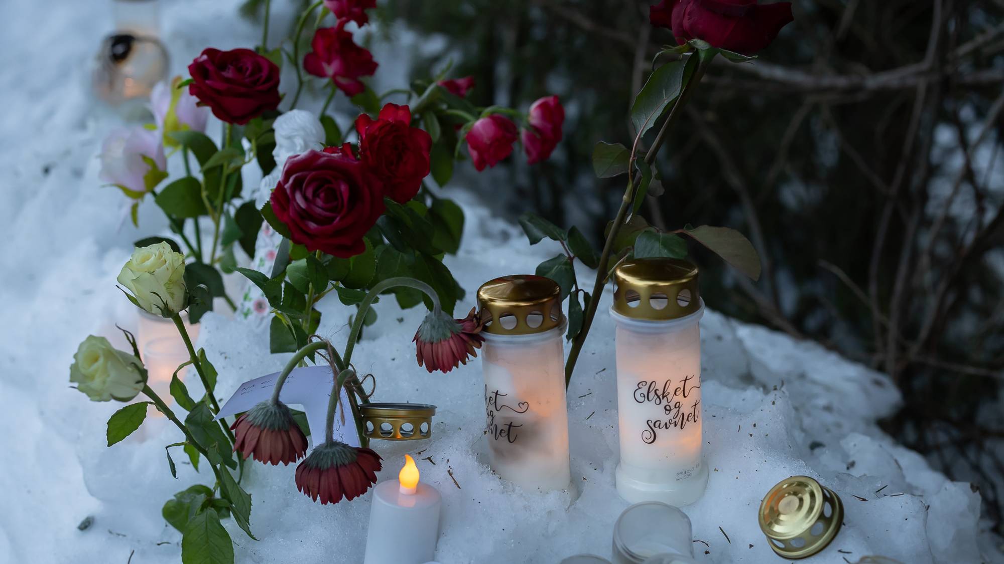 Bildet viser et minnesmerke med tennte lys og blomster som er plassert på snø. Røde roser og hvite blomster er lagt rundt flere lys med tekst på, som står på en snødekt overflate. Omgivelsene er preget av trær og natur, som gir kontekst til minnesmarkeringen. Dette bildet kan være knyttet til en sorgprosess eller en markering av en spesiell hendelse. (Bildebeskrivelsen er laget av en KI-tjeneste)