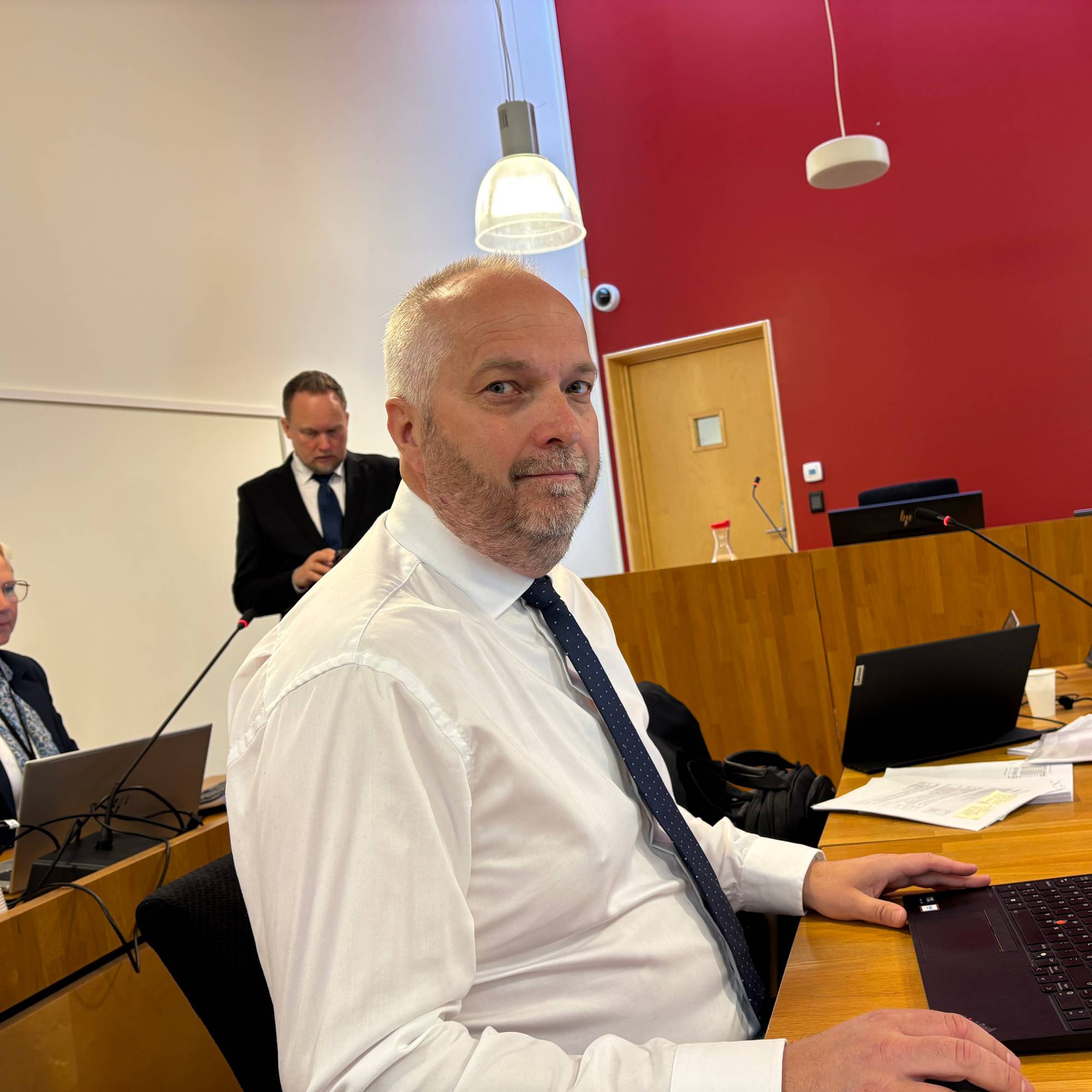 En mann i hvit skjorte med slips sitter ved et bord med en bærbar datamaskin. I bakgrunnen står en annen mann i dress. Til venstre sitter en kvinne. De befinner seg i et rom med rød vegg. (Bildebeskrivelsen er laget av en KI-tjeneste)