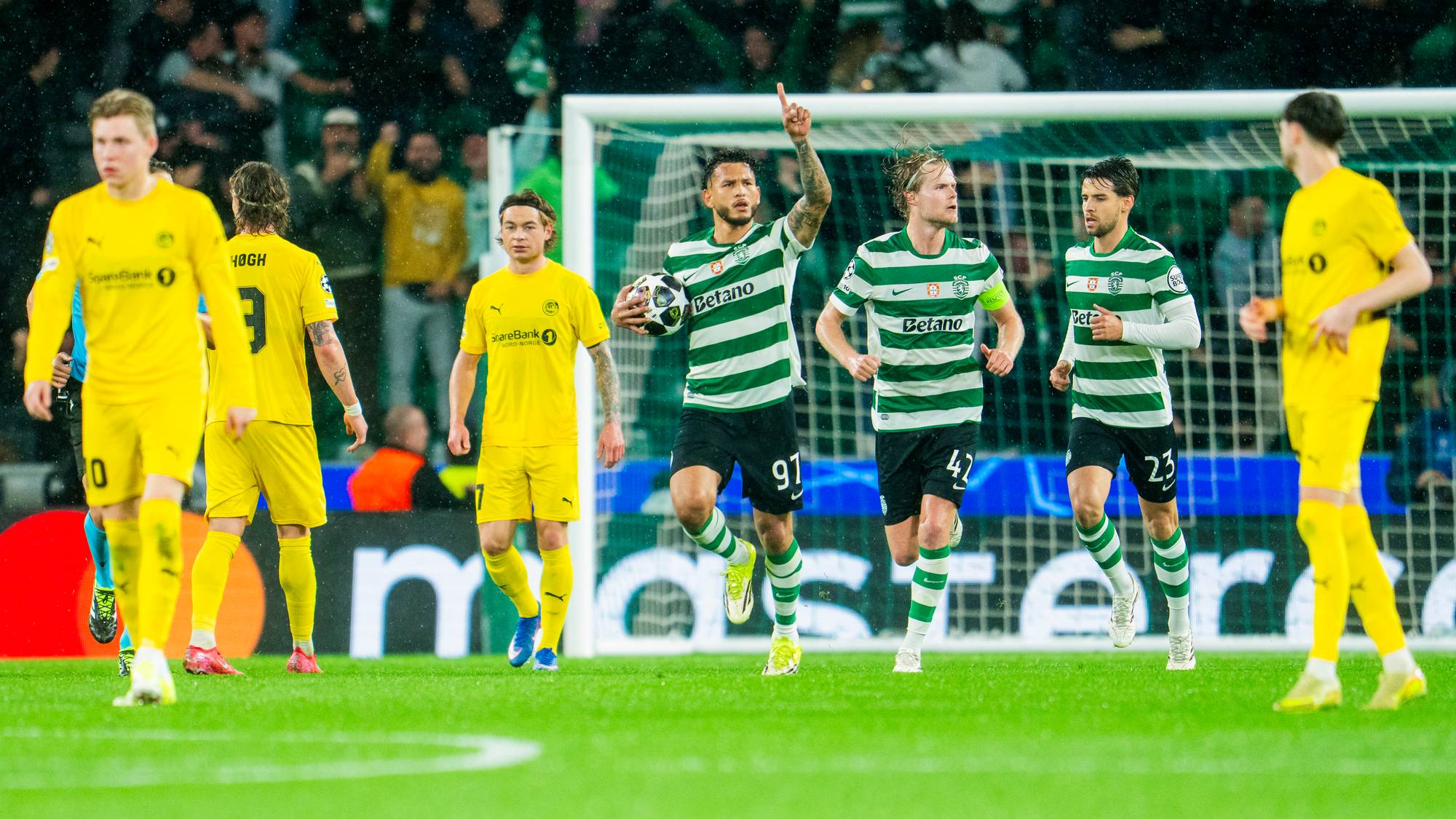 En spiller i grønn og hvit drakt med ball jubler og peker. Motspillere i gule drakter ser på. Publikum i bakgrunnen er uskarpt. (Bildebeskrivelsen er laget av en KI-tjeneste)