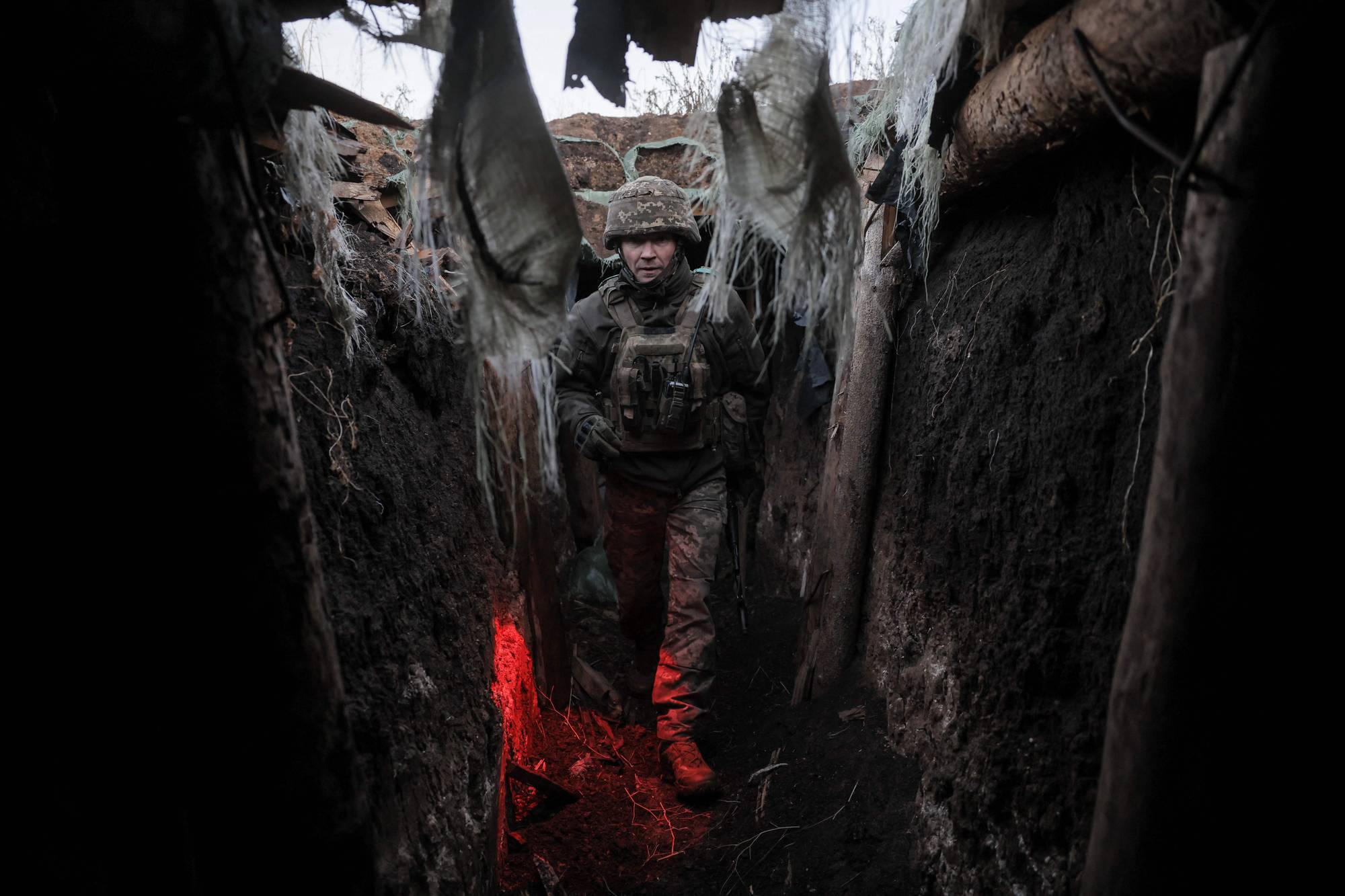 En soldat med hjelm og uniform går i en skyttergrav. Skyttergraven er mørk og har en rød lyskilde nederst i bildet. Bak soldaten er det synlig treverk og jordvegger. (Bildebeskrivelsen er laget av en KI-tjeneste)