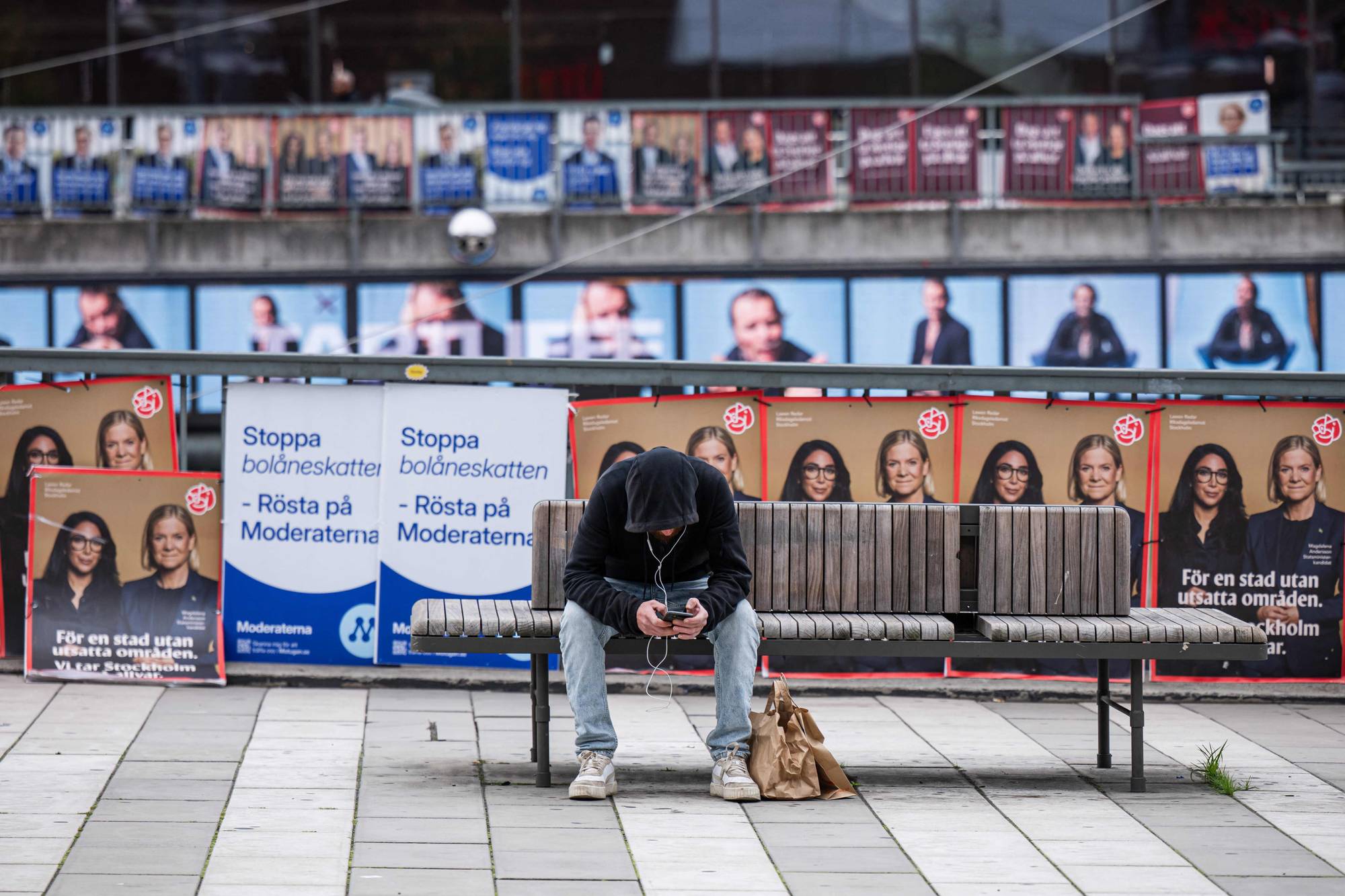 Person sitter på en benk og ser på mobil foran valgplakater for Moderaterna i Stockholm. (Bildebeskrivelsen er laget av en KI-tjeneste)
