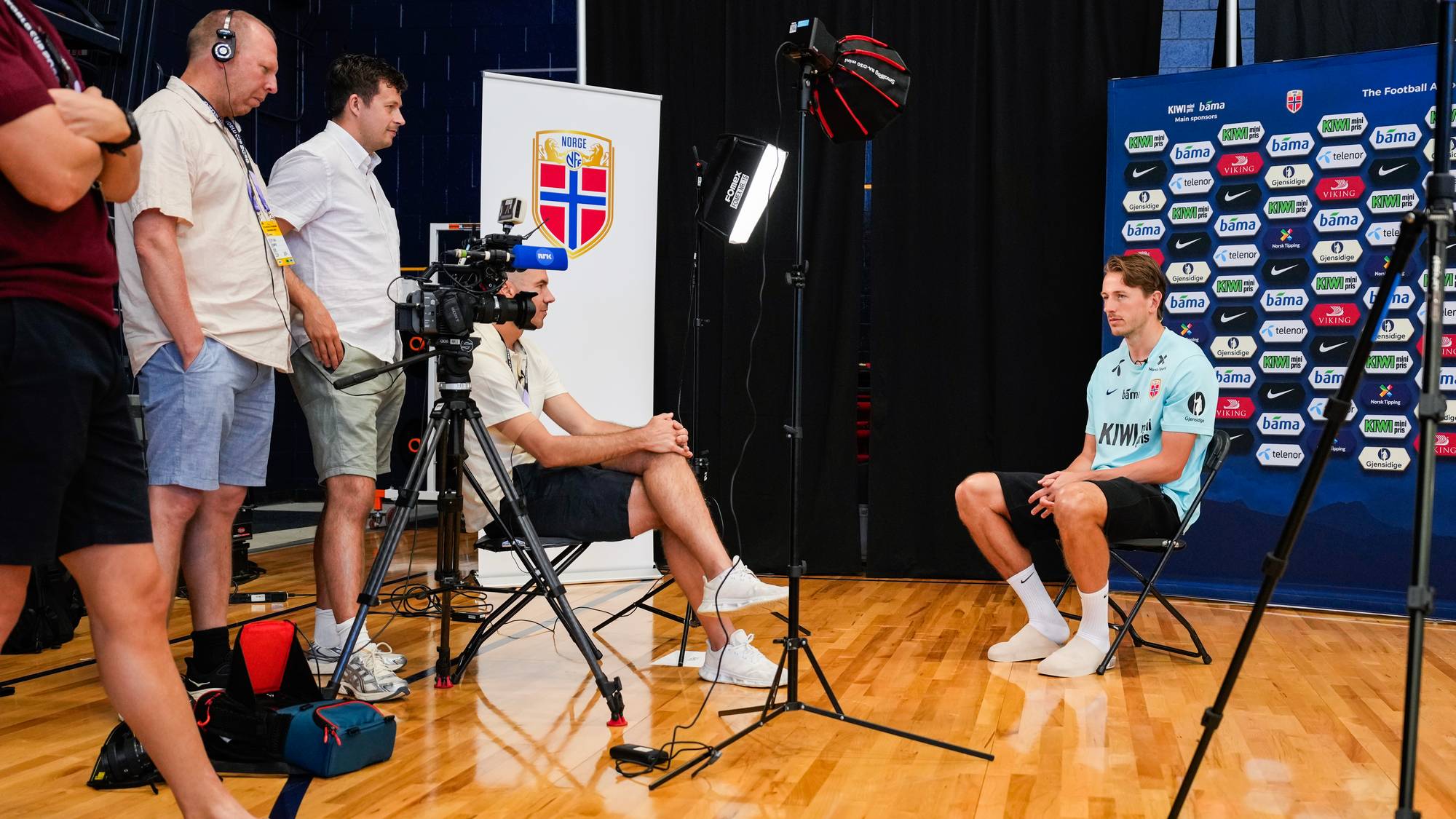 Fotballspiller sitter foran en sponsorvegg under et intervju. Ved siden av ham sitter en intervjuer, mens kameramenn og assistenter er synlige. Bakgrunnen består av sponsorlogoer og et banner med det norske flagget. (Bildebeskrivelsen er laget av en KI-tjeneste)