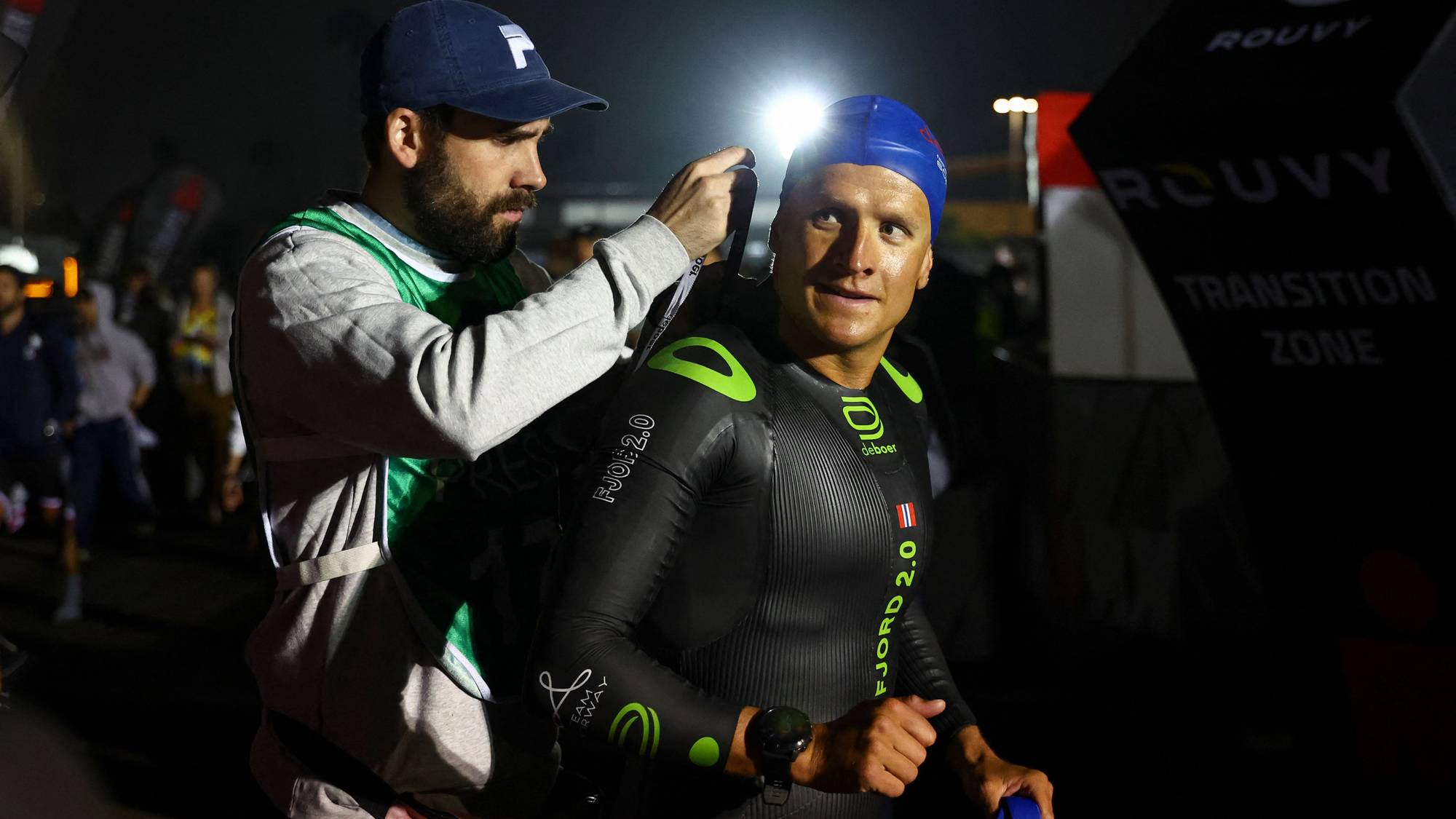 En mann i våtdrakt får hjelp av en annen mann. Mannen med våtdrakt har svømmehette og et norsk flagg på armen. Bak dem er det folk og en tavle med teksten «TRANSITION ZONE». (Bildebeskrivelsen er laget av en KI-tjeneste)