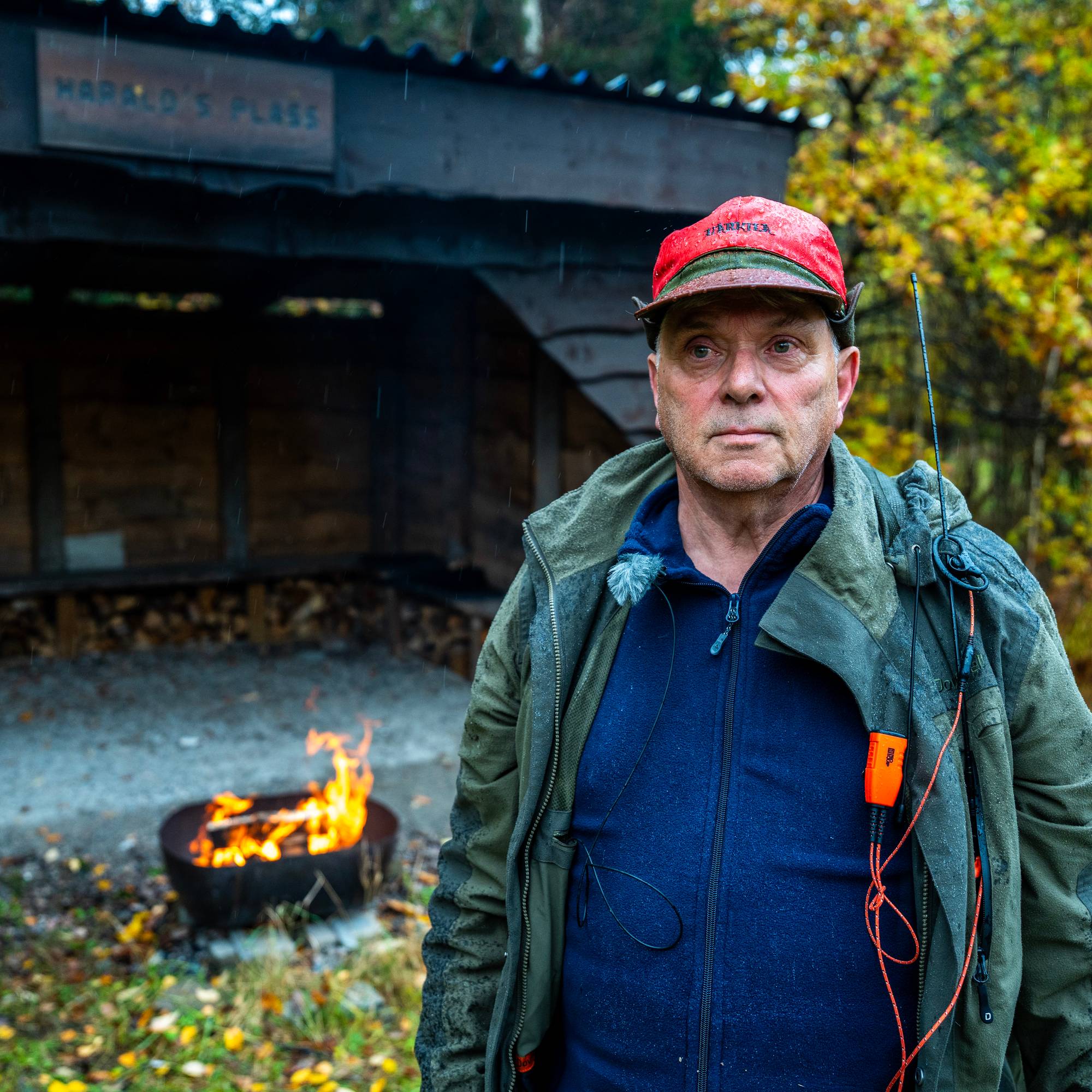 En eldre mann med rød caps står foran et bål ved en gapahuk merket Haralds plass. (Bildebeskrivelsen er laget av en KI-tjeneste)