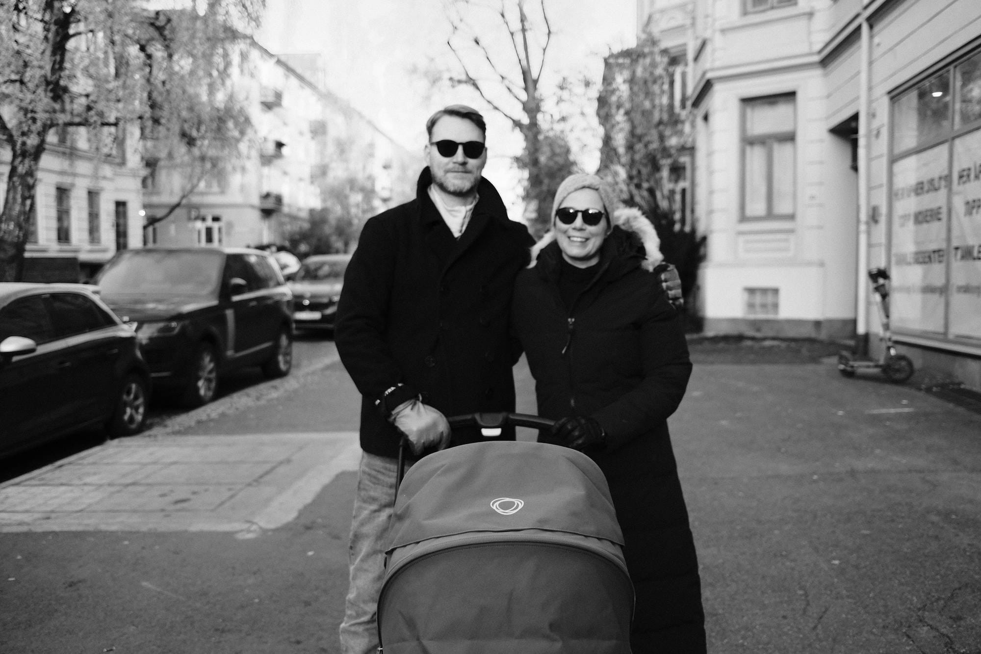 Mann og kvinne med solbriller står med en barnevogn i en bygate. Svart-hvitt fotografi.