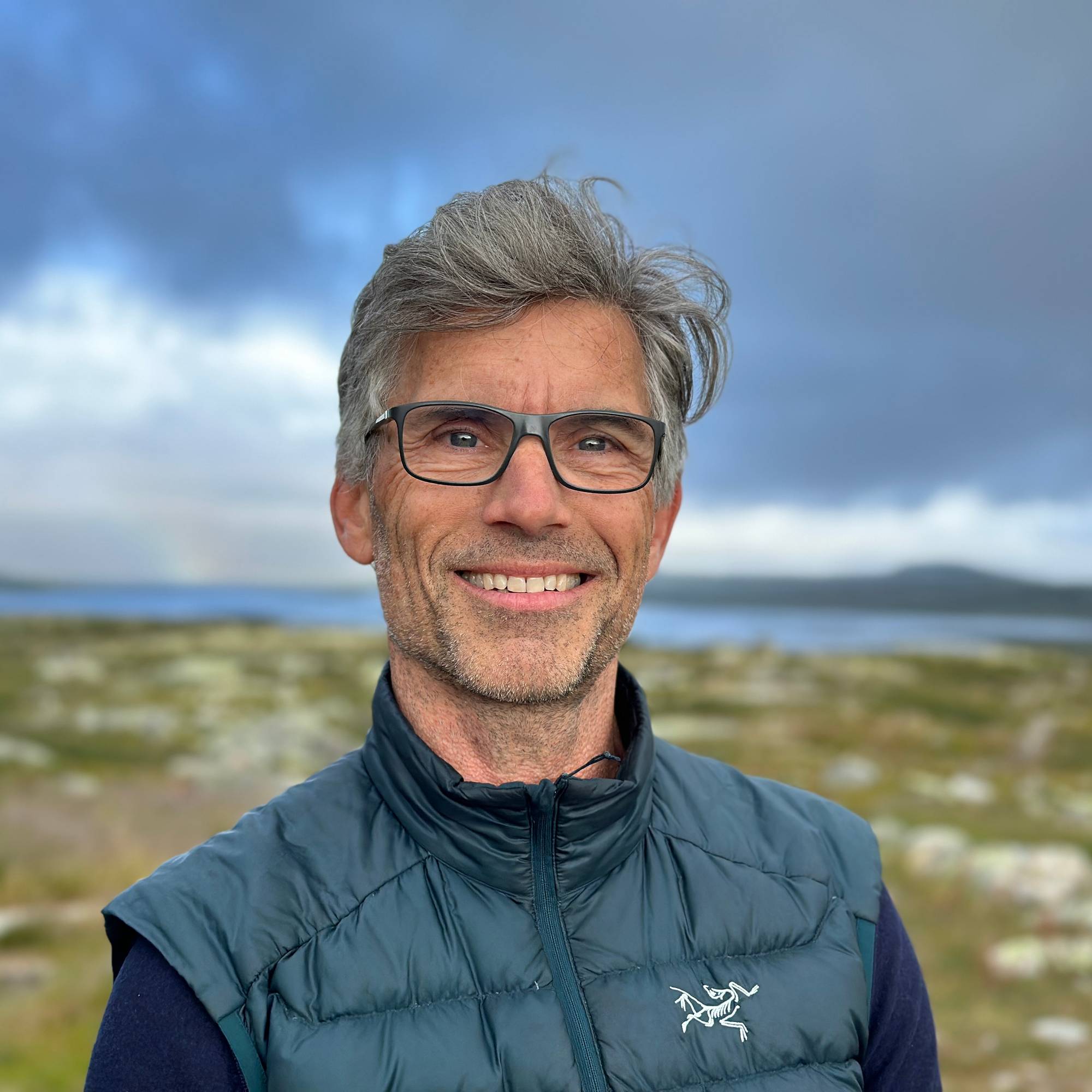 Mann med grått hår og briller foran grønn vidde og blått vann. 