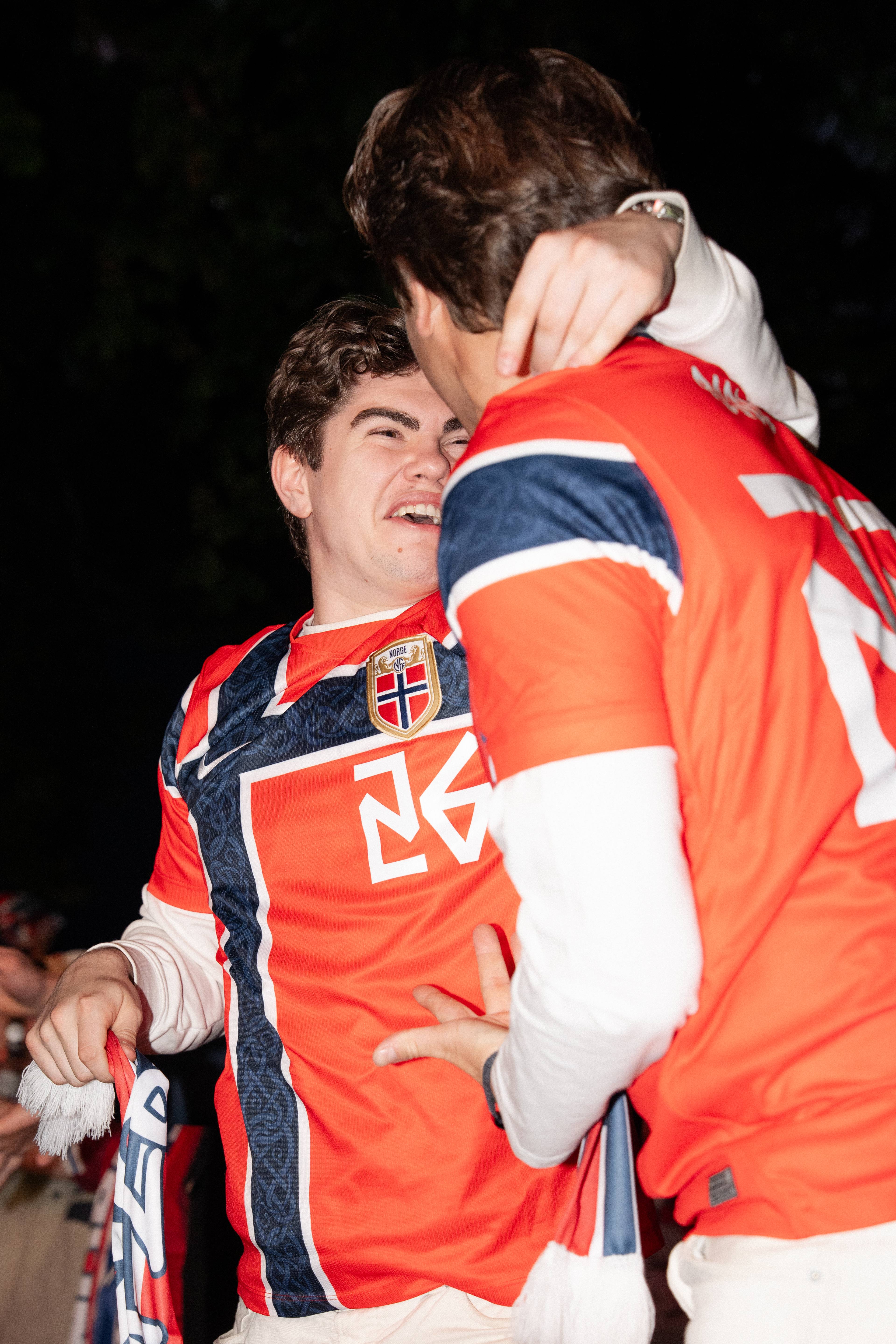 To unge menn i norske fotballdrakter. Den ene omfavner den andre mens han ler. Begge har på seg supporterutstyr. (Bildebeskrivelsen er laget av en KI-tjeneste)