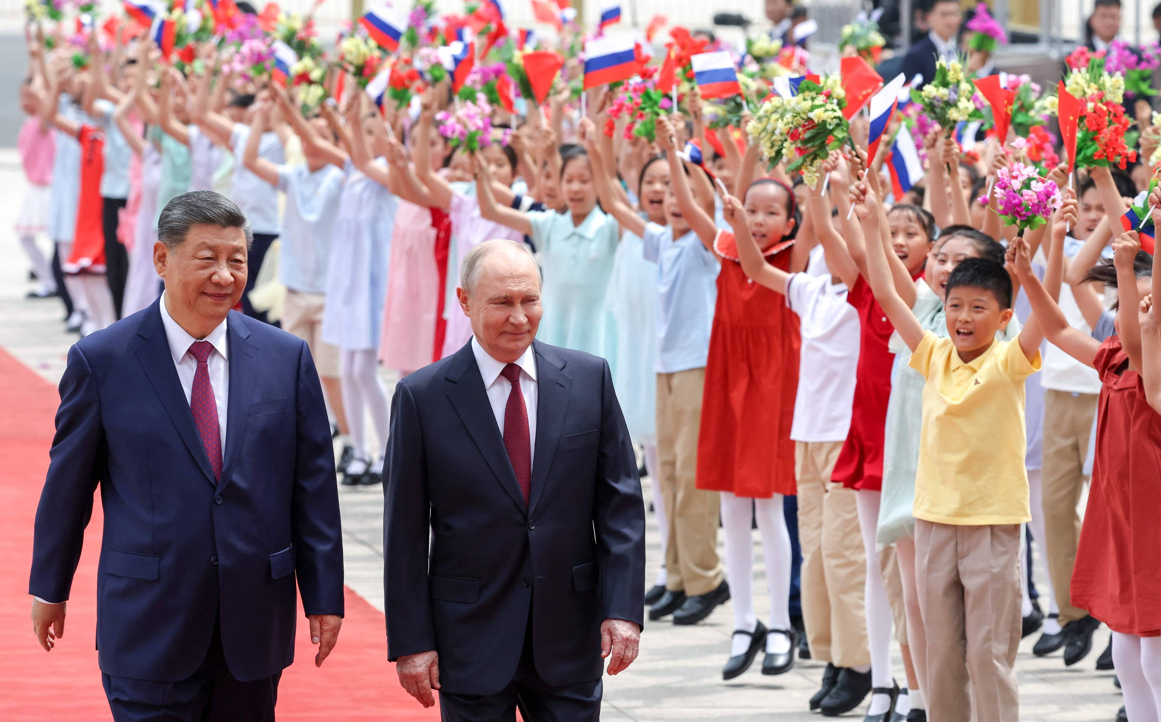 Kinas president Xi Jinping og Russlands president Vladimir Putin går på et rødt teppe. Bak dem vinker barn med blomster og flagg. Barnas opptreden skjer utendørs i en bygate. (Bildebeskrivelsen er laget av en KI-tjeneste)