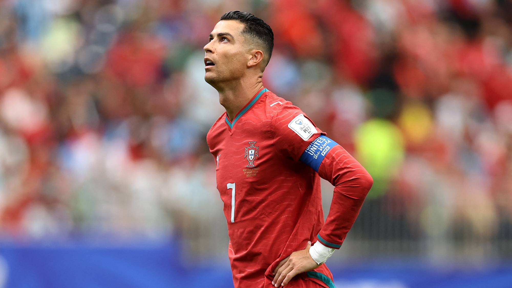 Fotballspiller Cristiano Ronaldo ser opp mens han står på banen. Han er ikledd en rød drakt med nummer 7 på brystet. I bakgrunnen sitter tilskuere på tribunen. (Bildebeskrivelsen er laget av en KI-tjeneste)