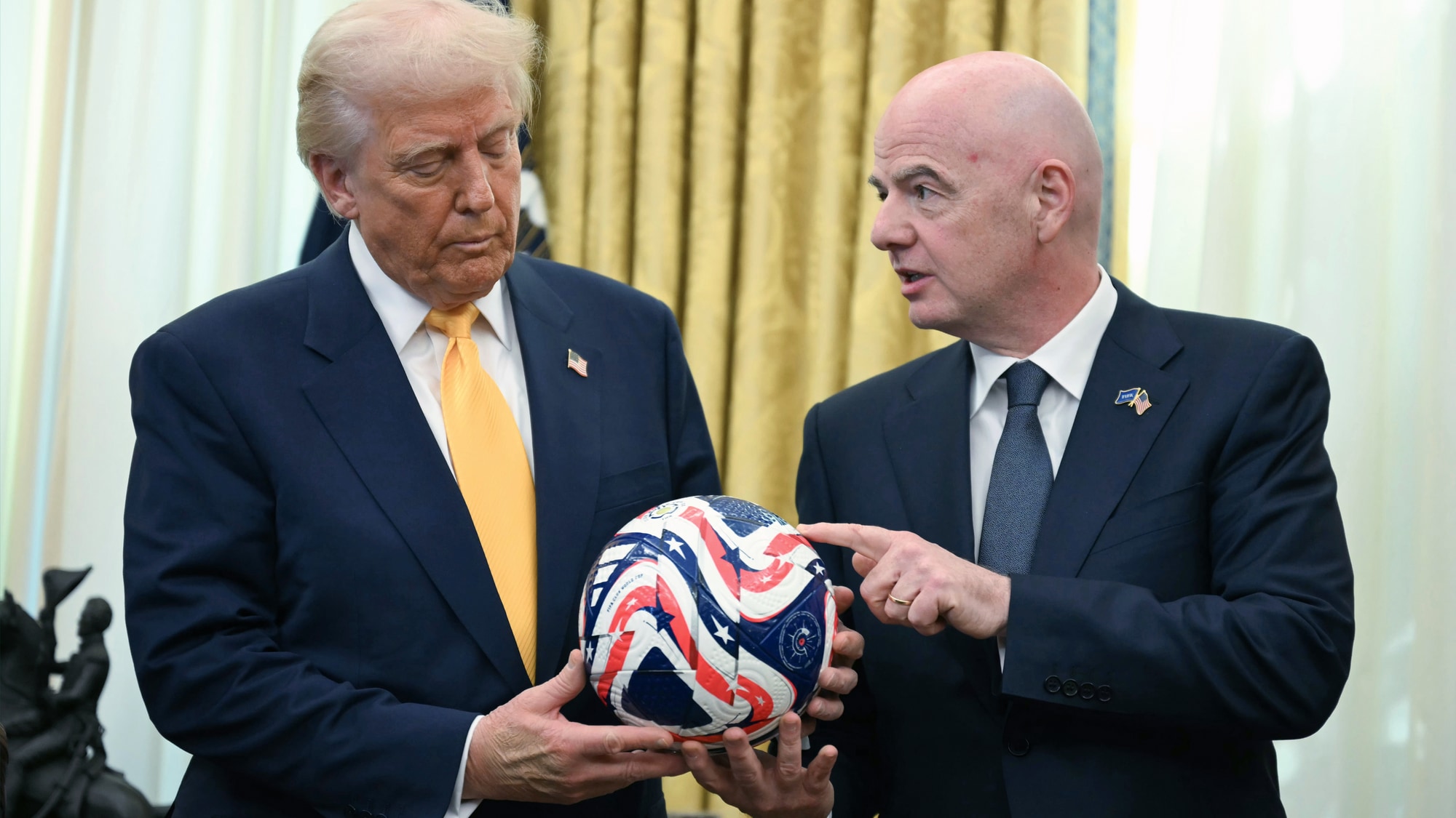 Donald Trump holder en fotball mens Gianni Infantino peker på den. Begge mennene er kledd i dress. De står innendørs foran et gyllent draperis. (Bildebeskrivelsen er laget av en KI-tjeneste)