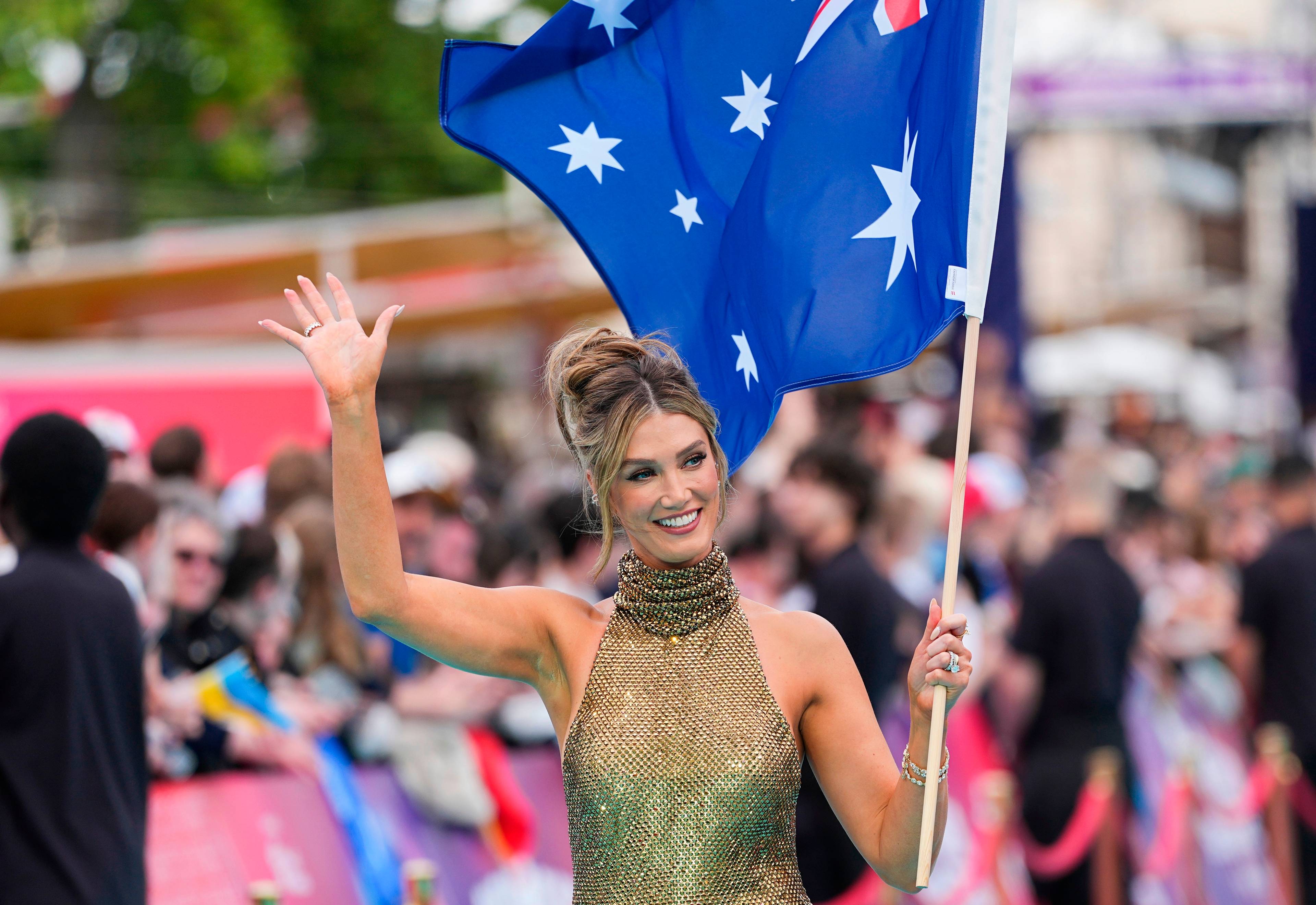 En kvinne vinker mens hun holder det australske flagget. Hun er kledd i en gullfarget kjole med paljetter. Kvinnen er omgitt av en folkemengde. (Bildebeskrivelsen er laget av en KI-tjeneste)