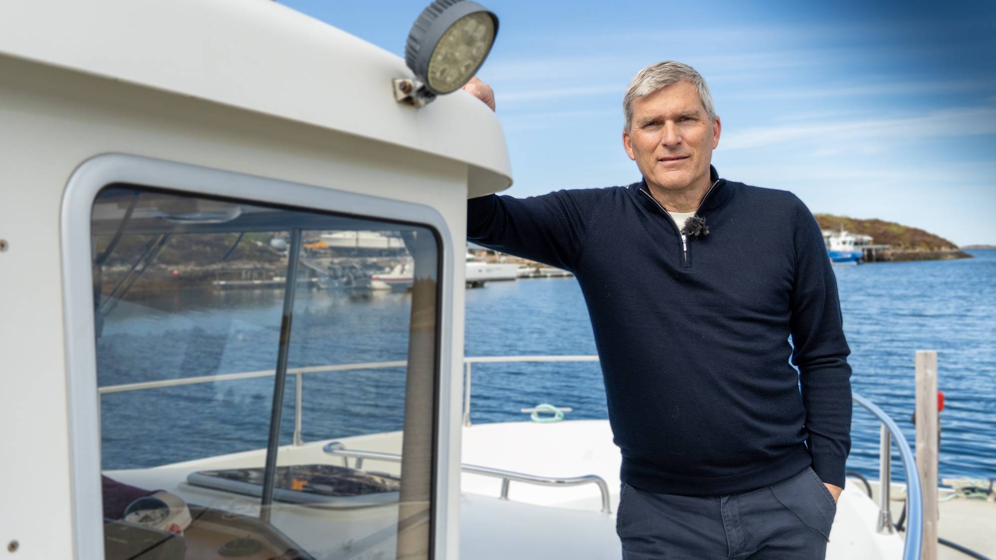 En mann står på en båt med en innsjø eller fjord i bakgrunnen. Mannen har grått hår og er kledd i en mørkeblå genser og bukser. Han ser mot kameraet og har en mikrofon festet til genseren. En lyskaster er montert på siden av båten. (Bildebeskrivelsen er laget av en KI-tjeneste)