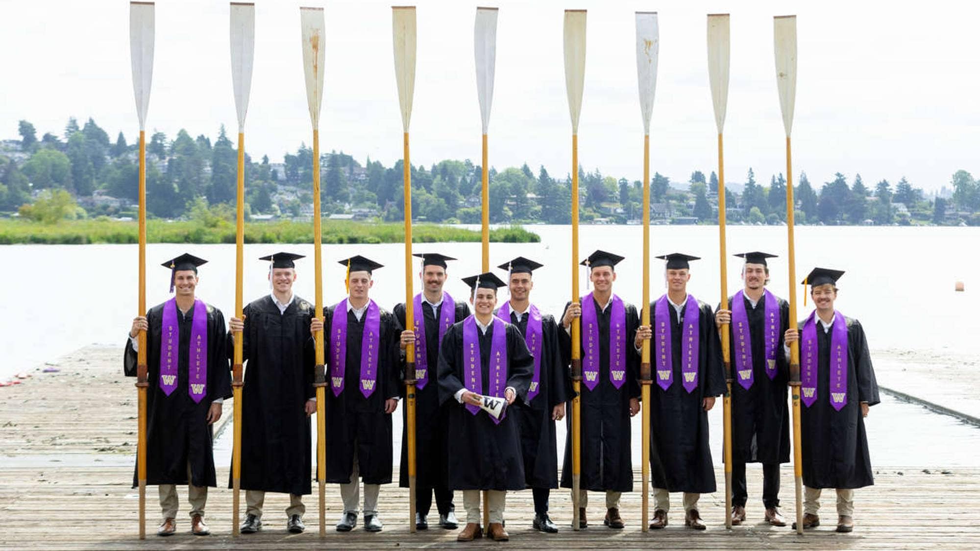 Ni mannlige studenter i akademisk kappe holder hver sin åre på en brygge ved et vann. (Bildebeskrivelsen er laget av en KI-tjeneste)