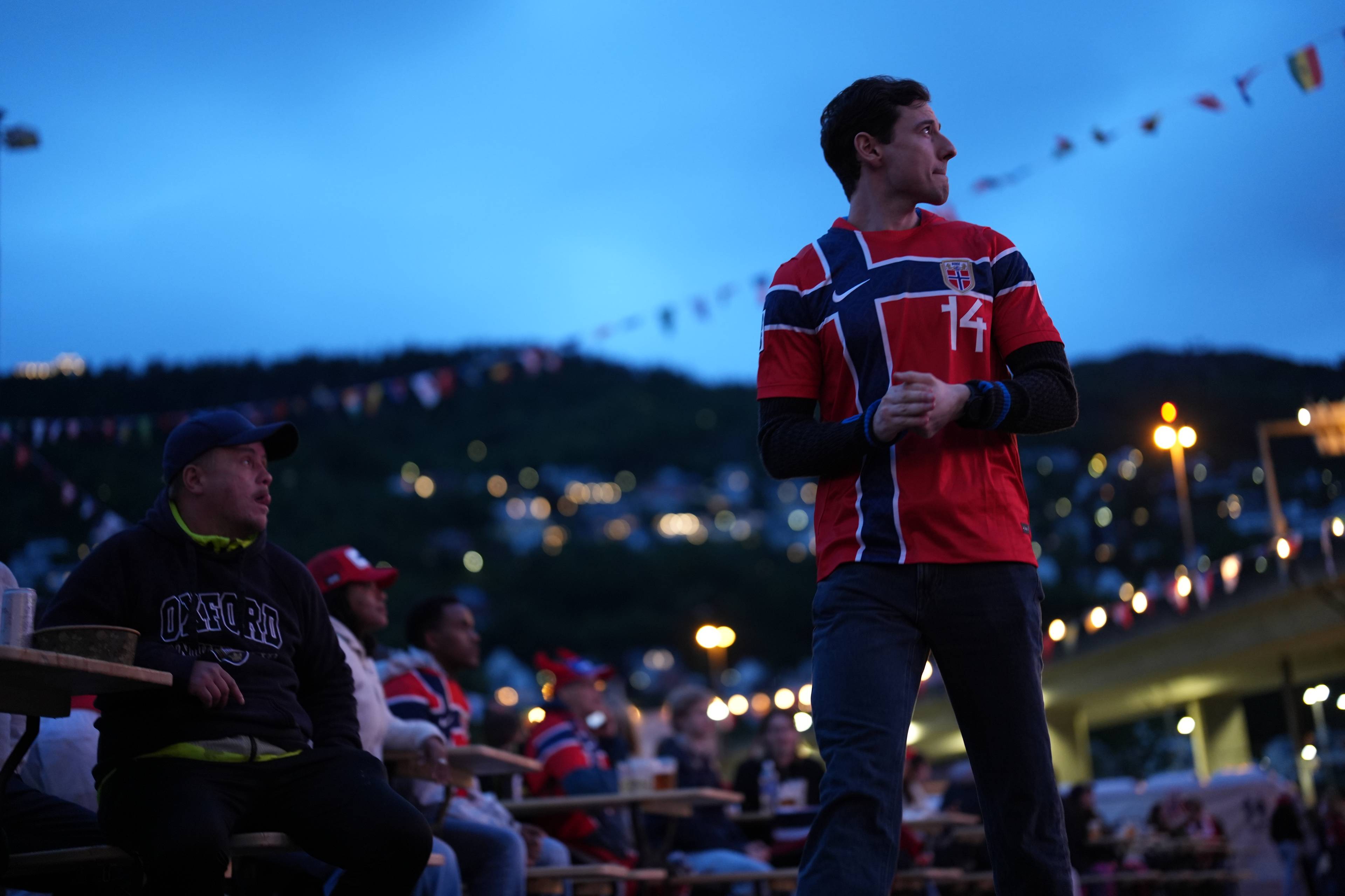 En mann i norsk fotballtrøye står og ser bort. Flere personer sitter ved bordene bak ham. Det er kveld og det henger flagg langs en åsside i bakgrunnen.